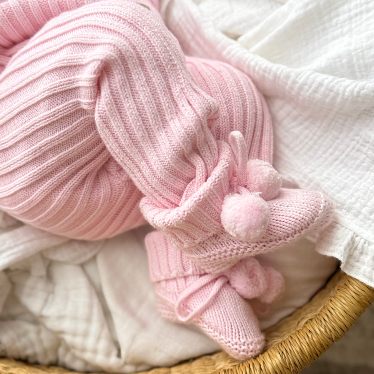 A close-up of a baby's legs wearing a pink ribbed knit romper and matching booties with pom-poms, resting in a woven basket lined with white muslin fabric.