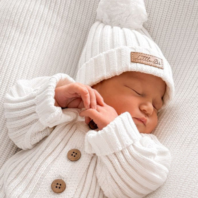 Newborn baby wearing a white ribbed knit romper and matching pom pom beanie with Little Bi branding