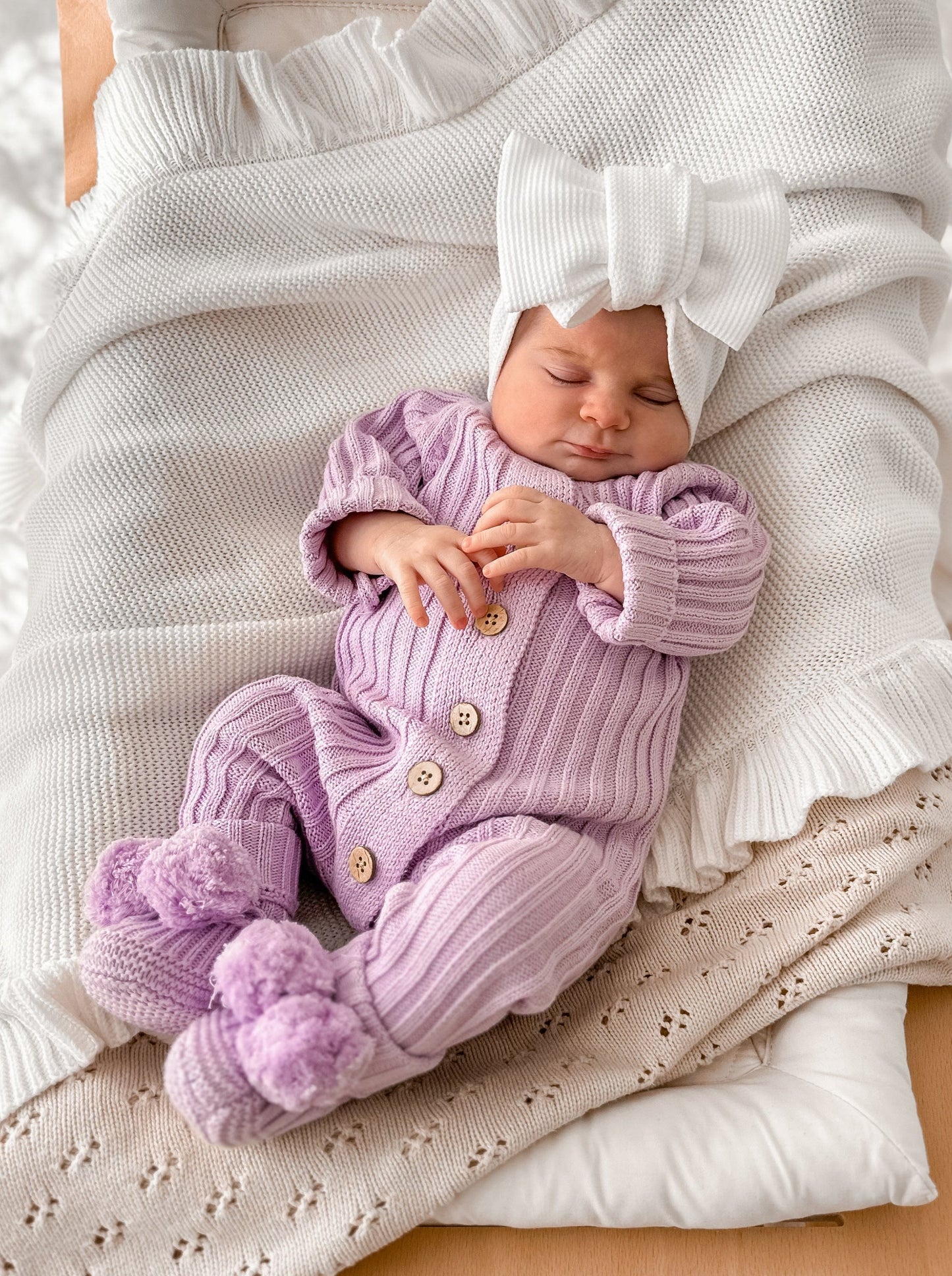 Baby wearing purple cotton romper and matching purple booties with pom poms and oversized white bow