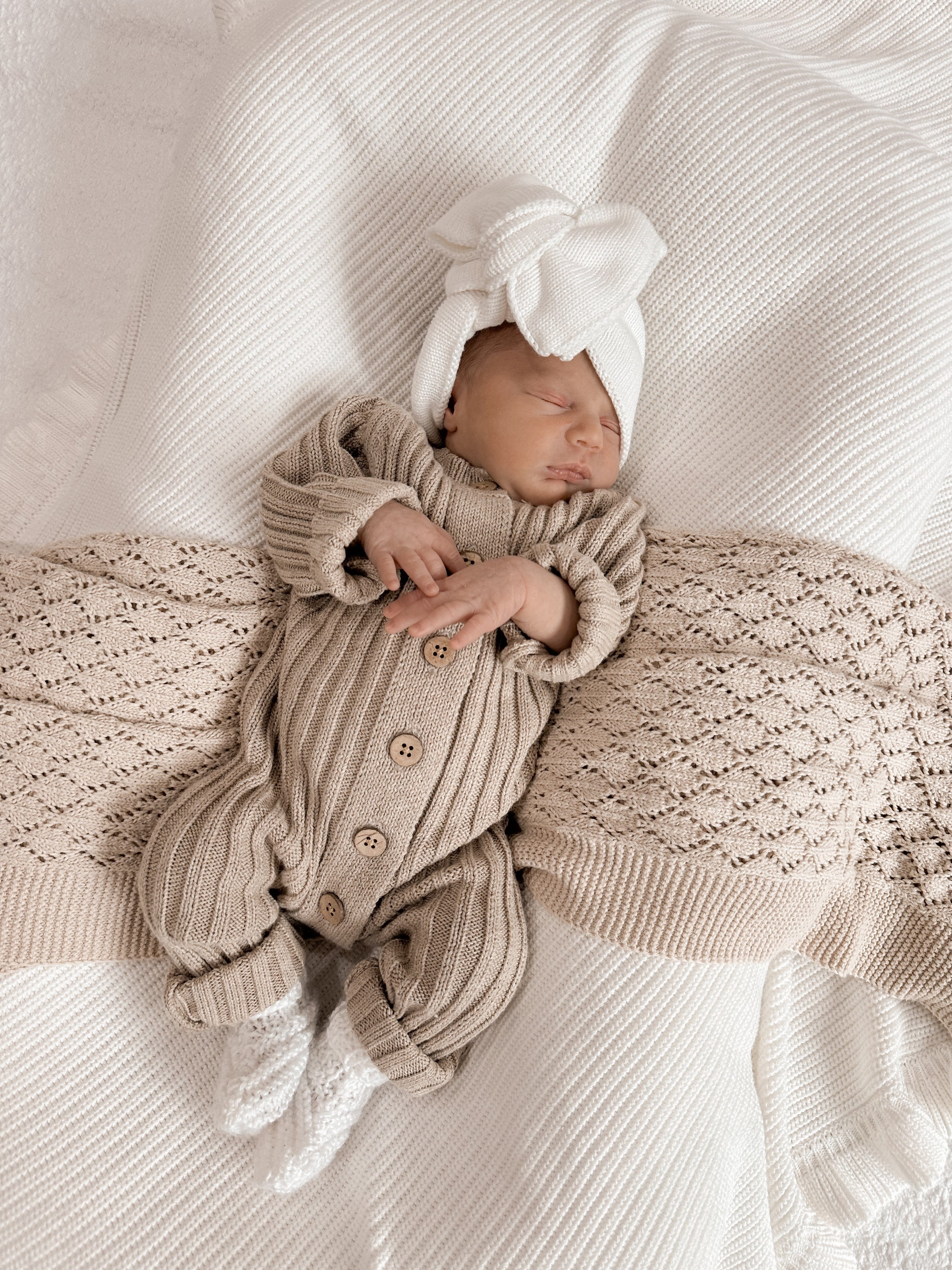 Newborn baby sleeping in a Little B's Retail Knitted Cotton Ribbed Romper - Oak with a white bow hat