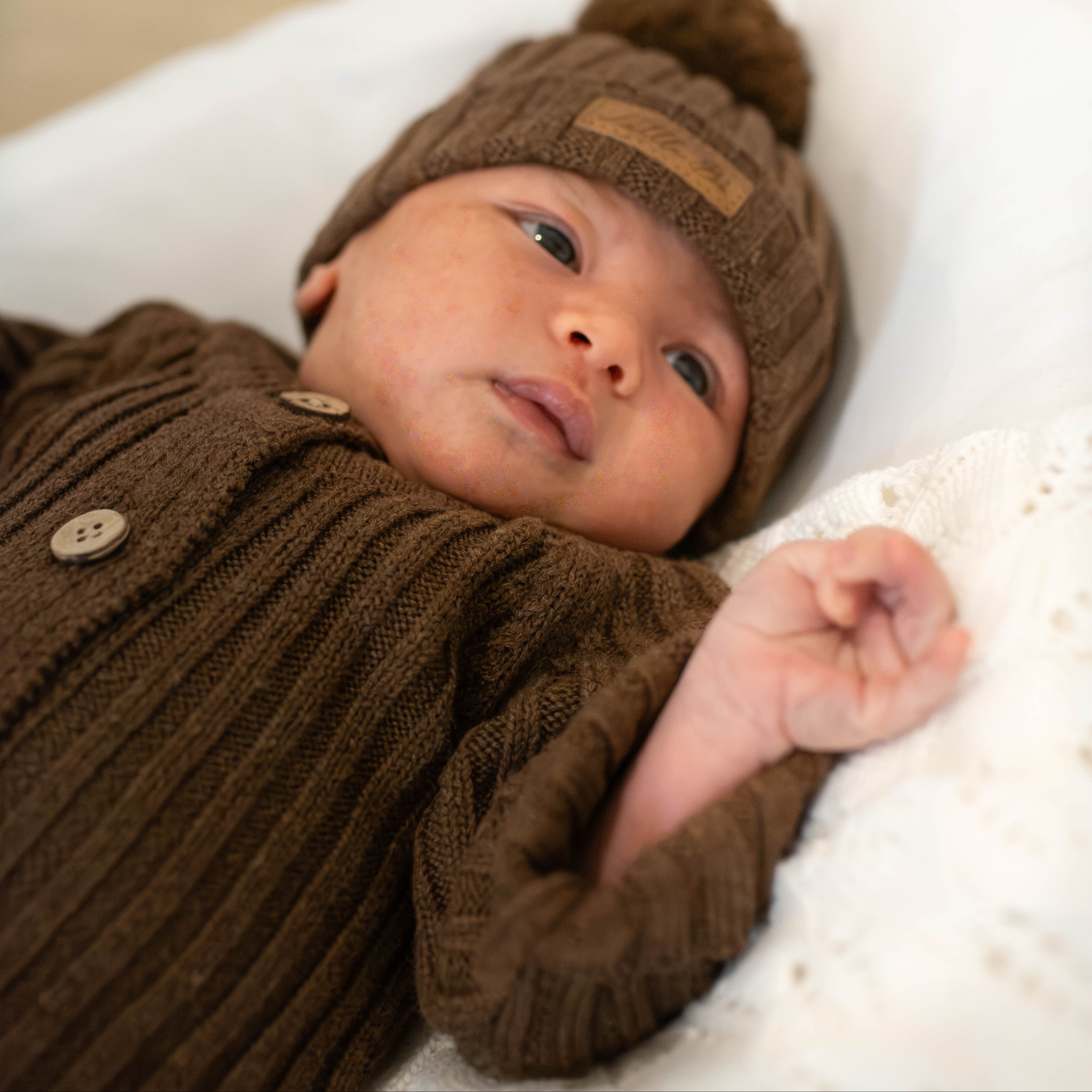 Baby laying on neutral background wearing knitted ribbed romper and matching beanie in chocolate brown