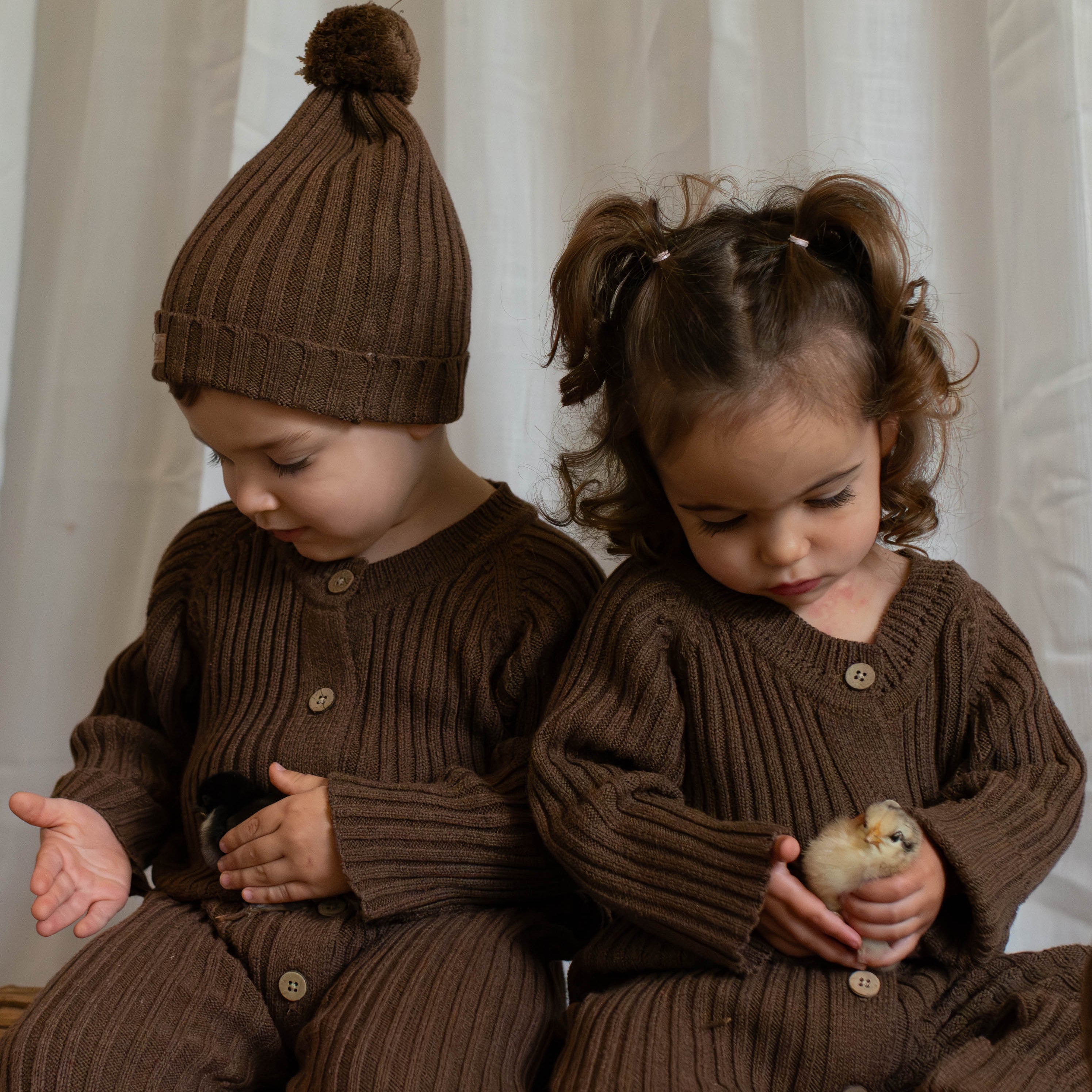 A boy and girl toddlers holding baby chickens and wearing chocolate brown knitted ribbed rompers