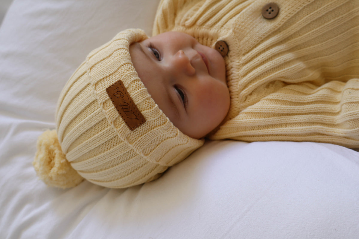 A baby lying on white bedding, wearing a yellow-coloured ribbed knit romper and matching pom-pom beanie with a brown leather brand patch.