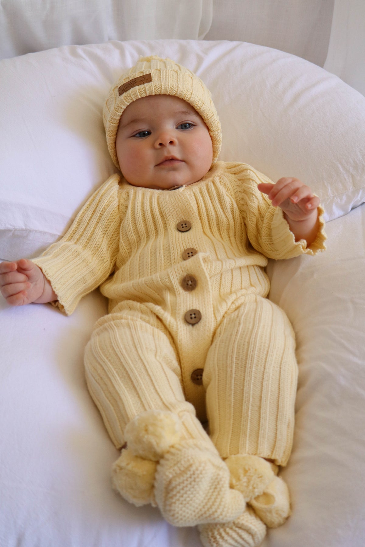 A baby lying on a white pillow, wearing a pale yellow ribbed knit one-piece outfit with matching booties and hat, featuring brown buttons down the front.