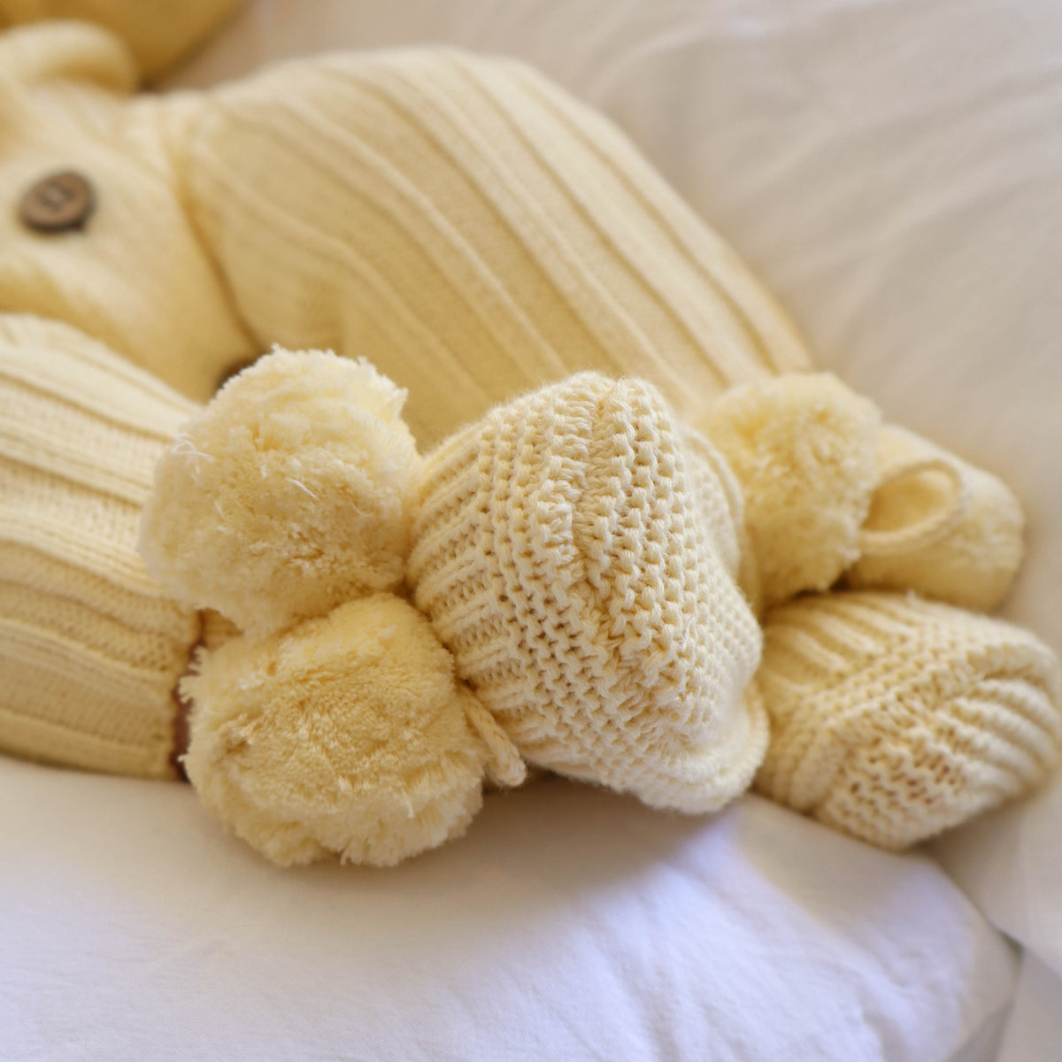 A close-up of a soft yellow ribbed knit baby outfit with matching pom-pom booties resting on white bedding.