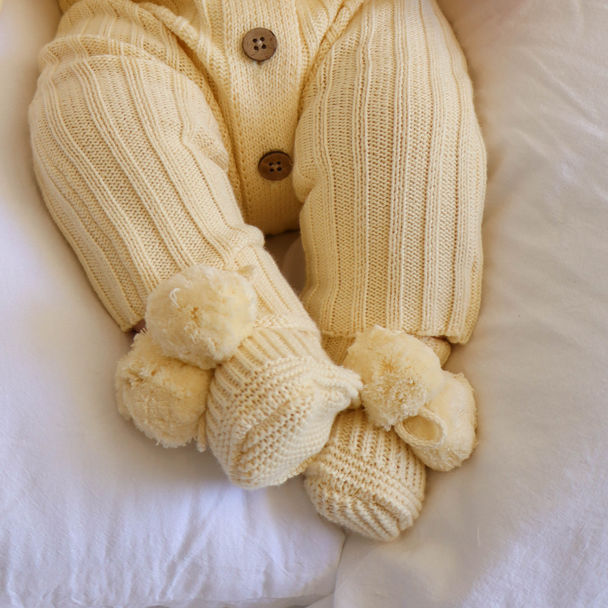 A close-up of a baby's legs wearing a cream-colored ribbed knit cardigan and matching booties with fluffy pom-poms, resting on white bedding.