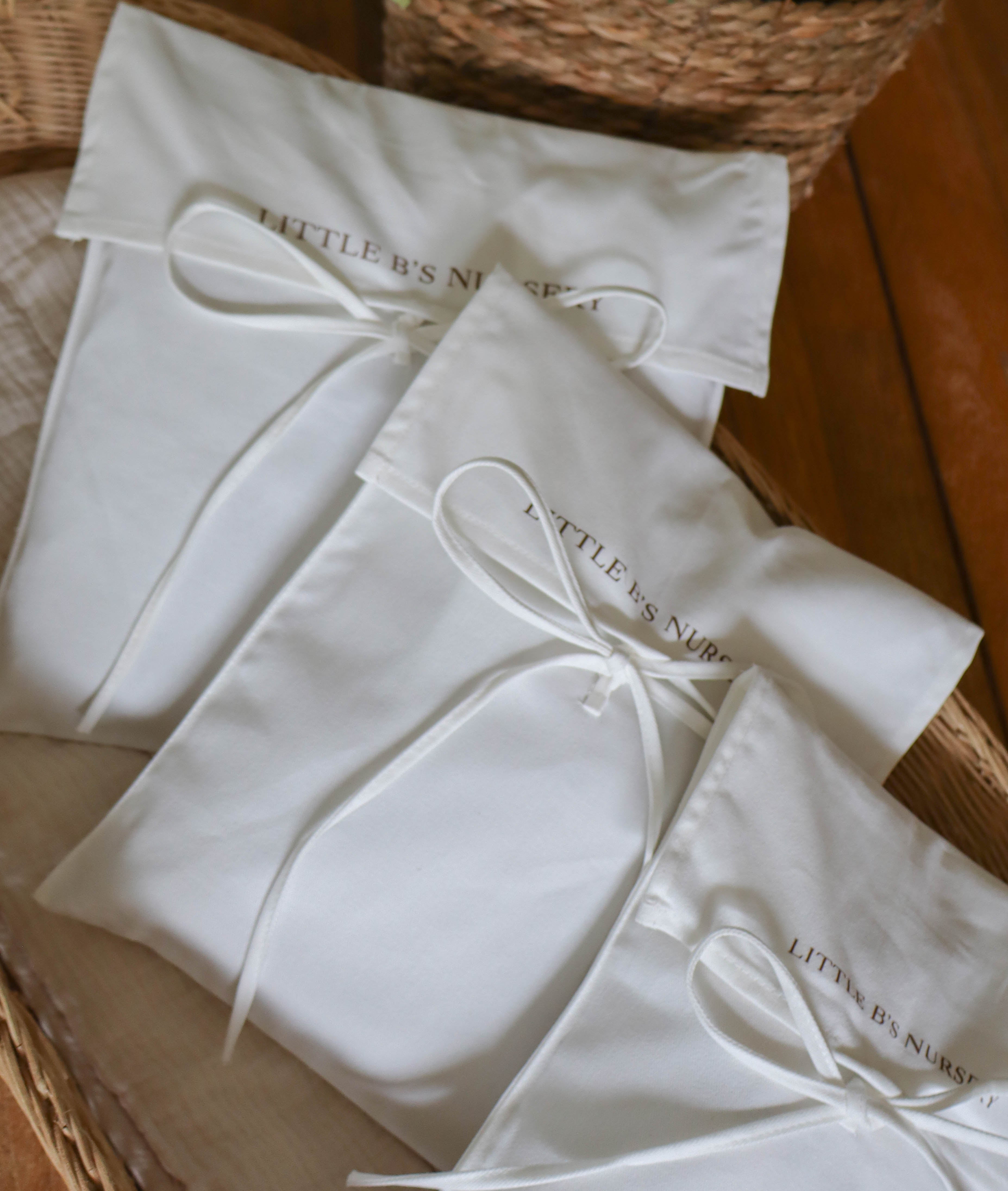 Three white fabric gift pouches with white drawstrings and gold foil text reading 'LITTLE B'S NURSERY' arranged in a woven basket.