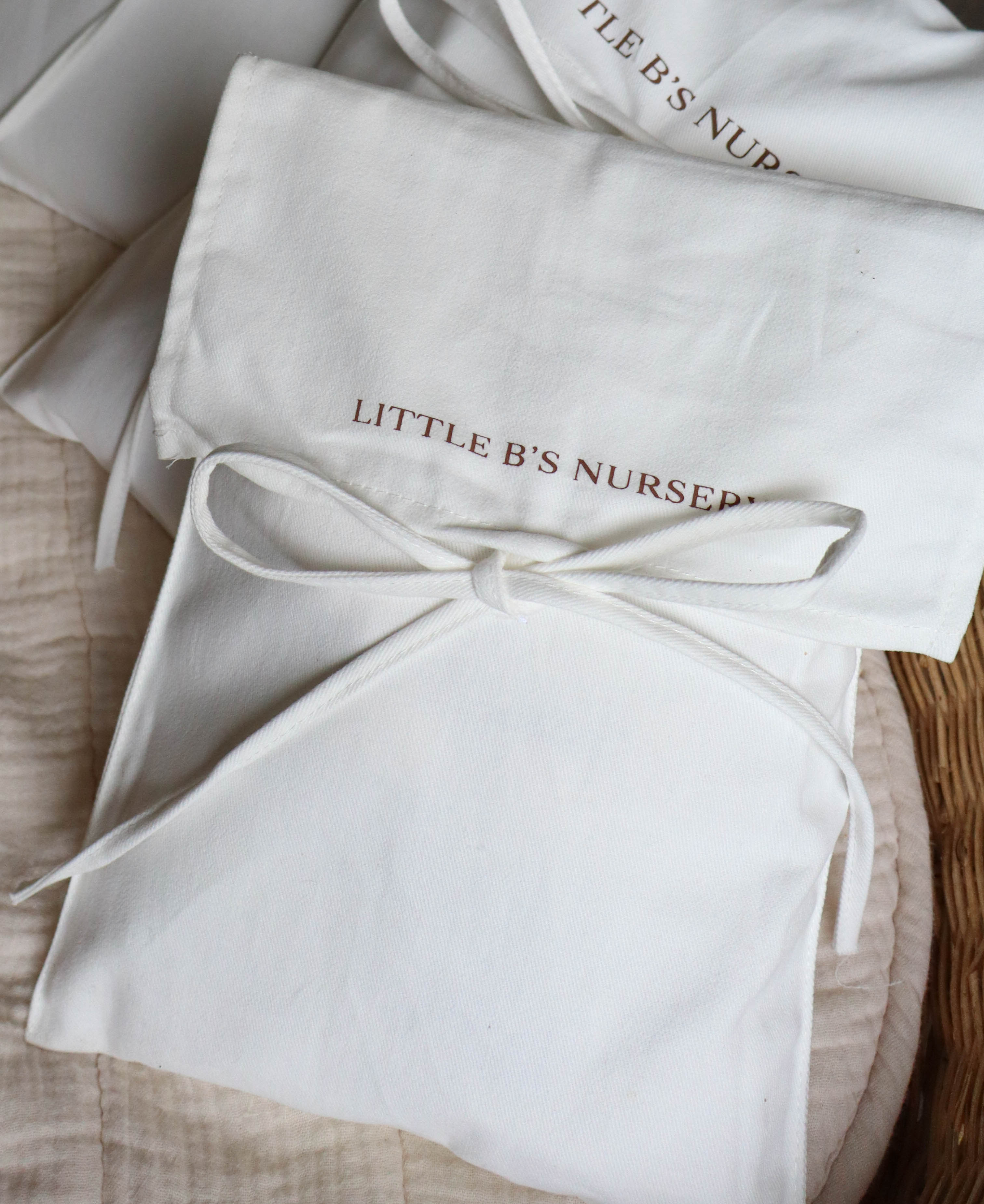 A stack of white fabric gift bags tied with white strings, printed with the words 'LITTLE B'S NURSERY' in a warm brown tone, arranged on a textured beige fabric surface.