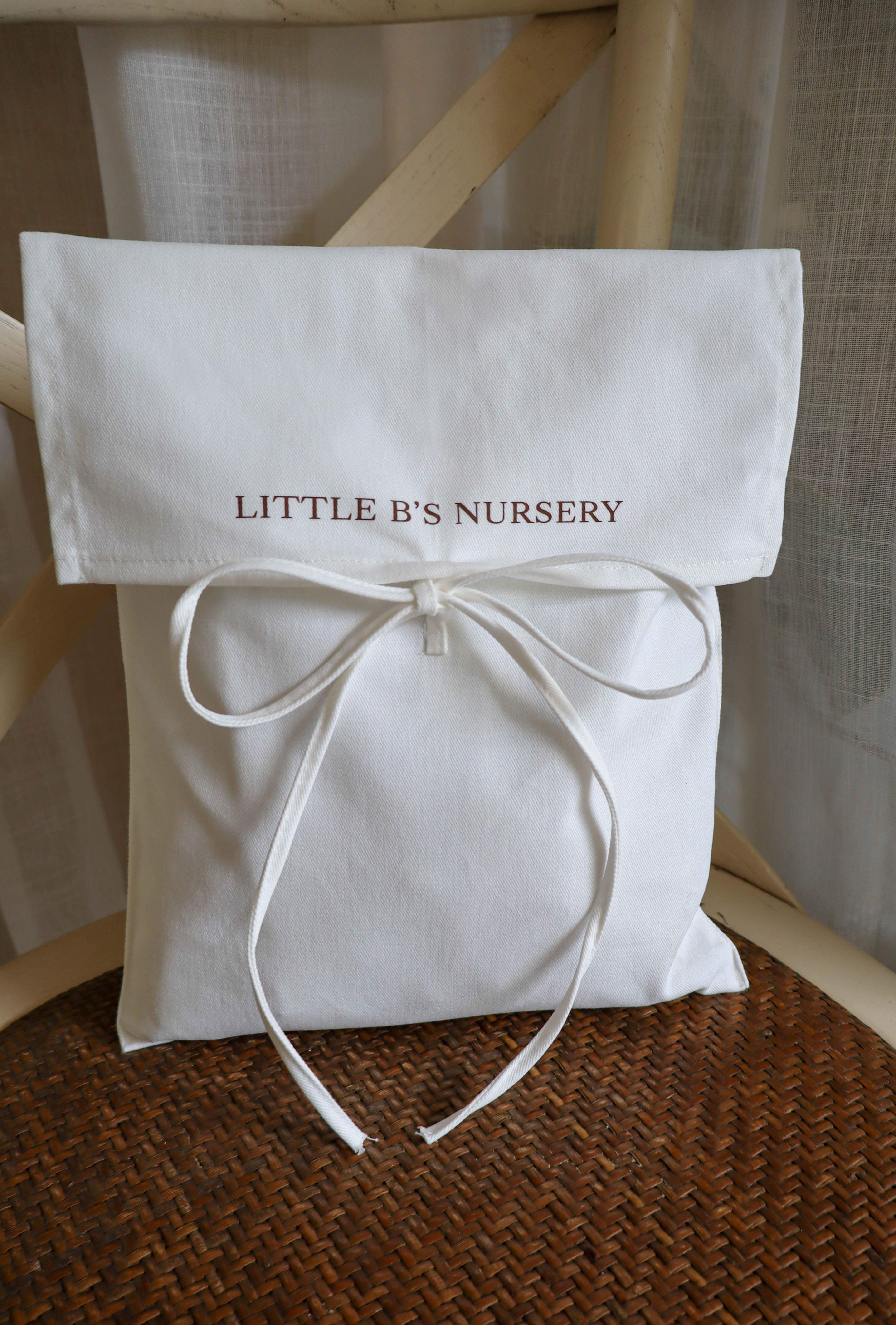 A white fabric nursery gift bag or pouch with a rolled top flap, embroidered with 'LITTLE B'S NURSERY' in brown, tied with a matching ribbon bow. The bag is resting on a brown woven rattan surface in front of a light wood chair and white curtain background.