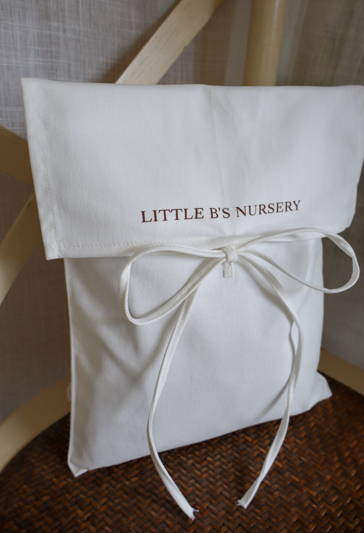 A white fabric pouch or gift bag with a fold-over flap printed with the words 'LITTLE B'S NURSERY' in brown lettering, tied with a matching white ribbon bow. The bag is placed on a brown woven surface with a light wood chair visible in the background.