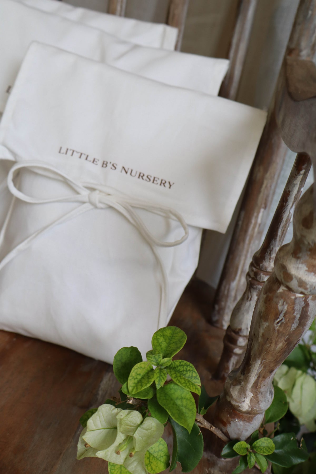 A cream-colored fabric gift bag with the words 'LITTLE B'S NURSERY' printed in gold, tied with a matching cream ribbon. The bag is styled on a rustic wooden surface next to green plants.