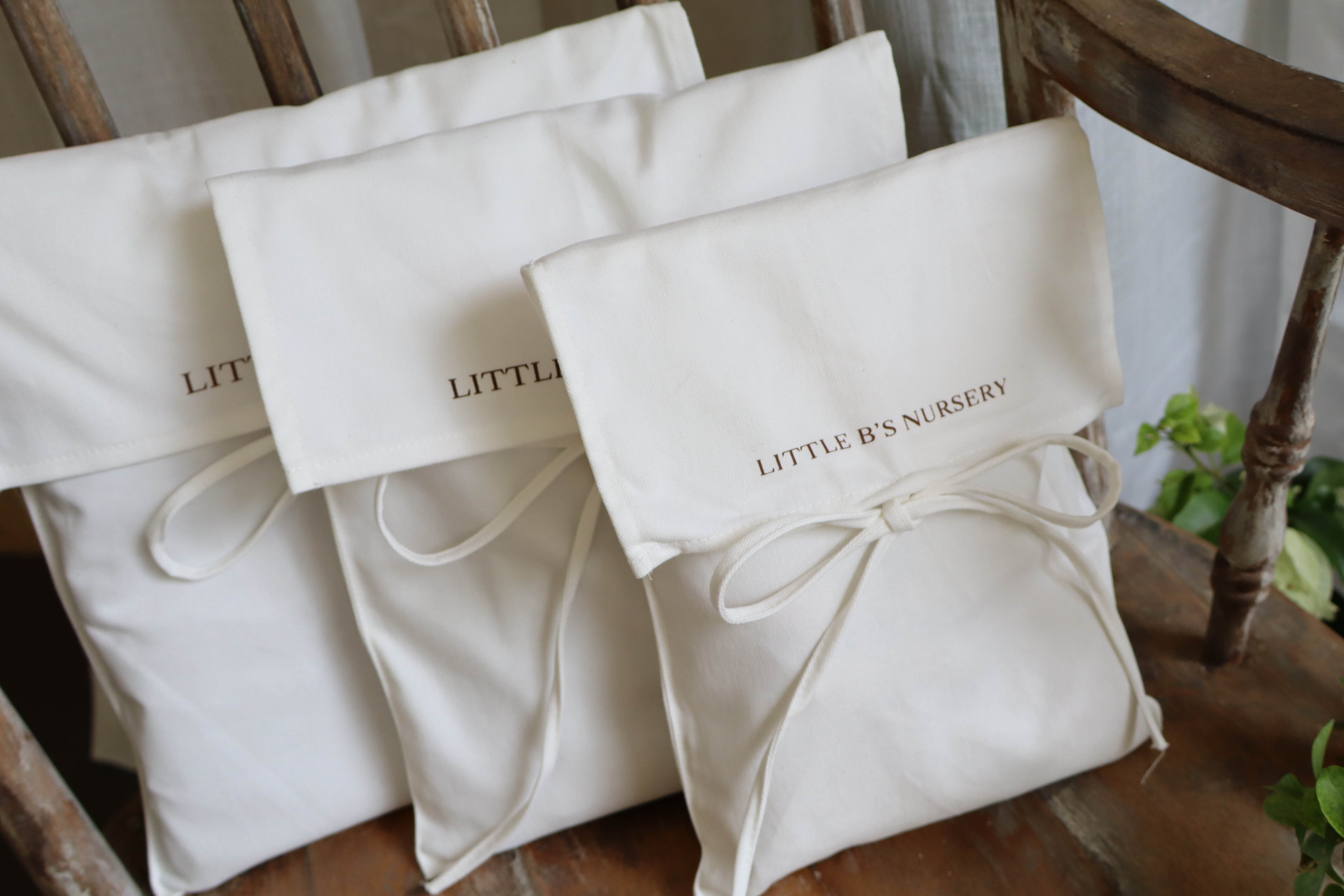 Three cream-colored fabric pouches with drawstring ties and embroidered 'LITTLE B'S NURSERY' logos, arranged on a rustic wooden chair.