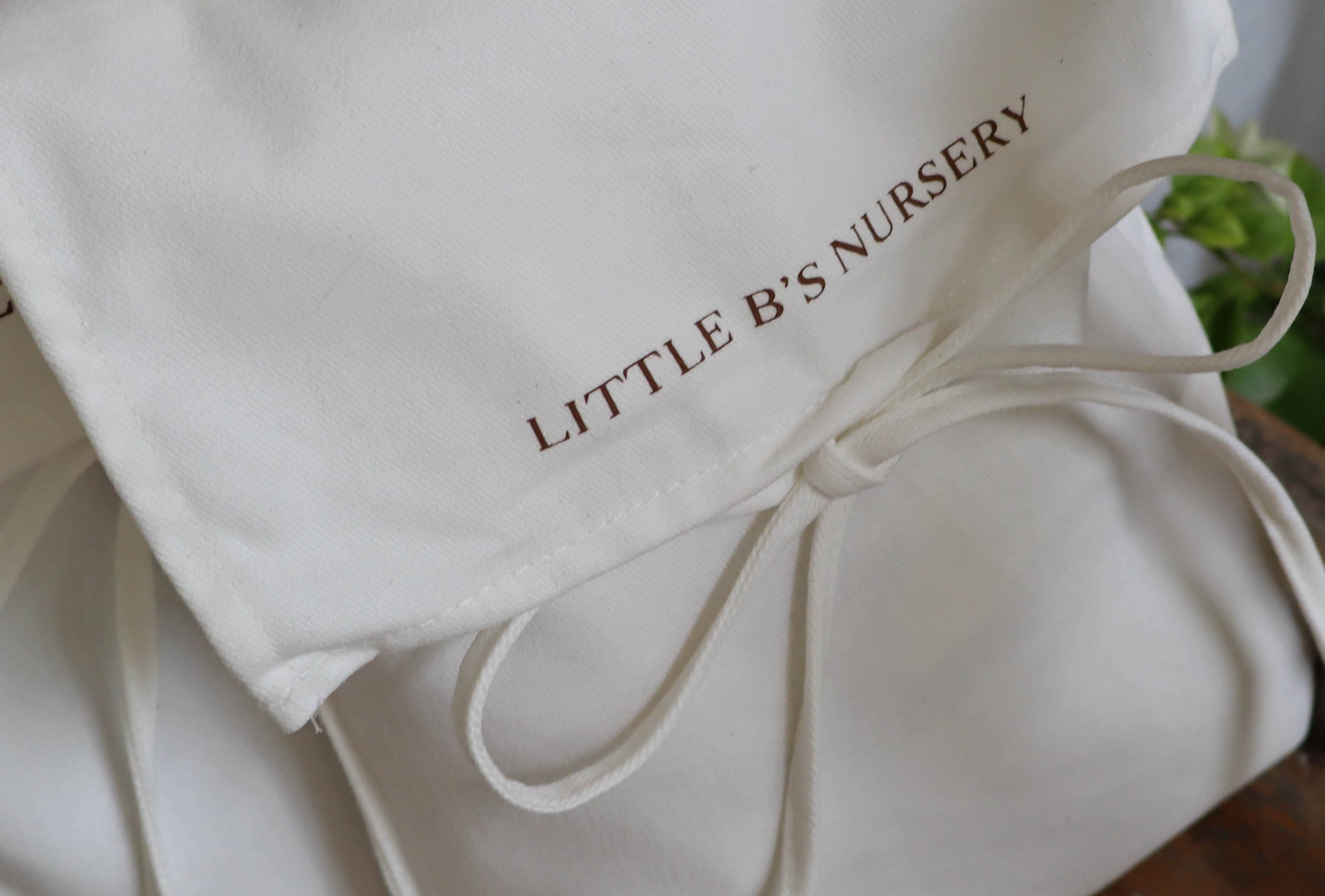 A close-up of a white fabric nursery gift bag or pouch with the words 'LITTLE B'S NURSERY' printed in brown on the front, tied with a matching white cord.