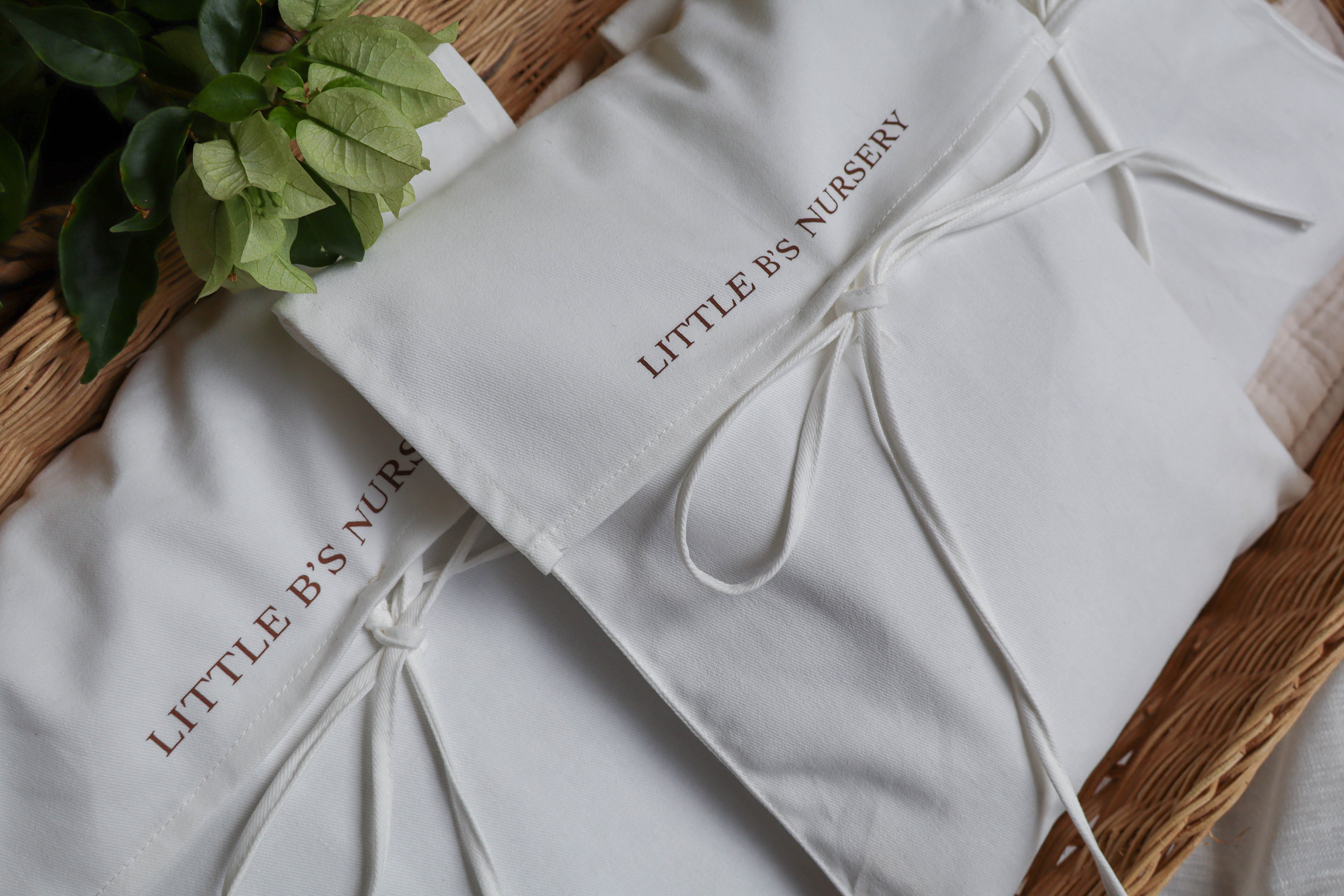 Two white fabric baby nursery gift bags with drawstring ties, printed with 'LITTLE B'S NURSERY' in brown lettering, arranged on a wicker tray with green leaves in the corner.