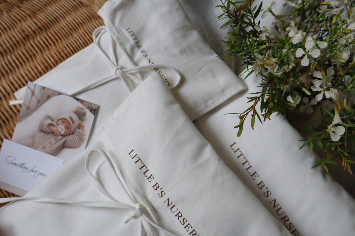 A styled flat-lay of white baby bedding or fabric accessories with the words 'LITTLE BIS NURSERY' printed in gold, arranged on a woven mat with a small photo card and floral accents.