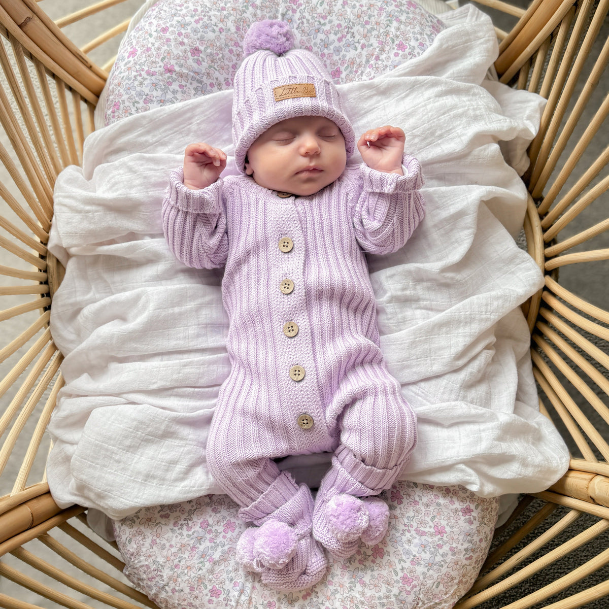 A sleeping newborn baby wearing a light purple ribbed knit one-piece outfit with matching pom-pom hat and footies, lying on a white muslin sheet inside a wicker basket.