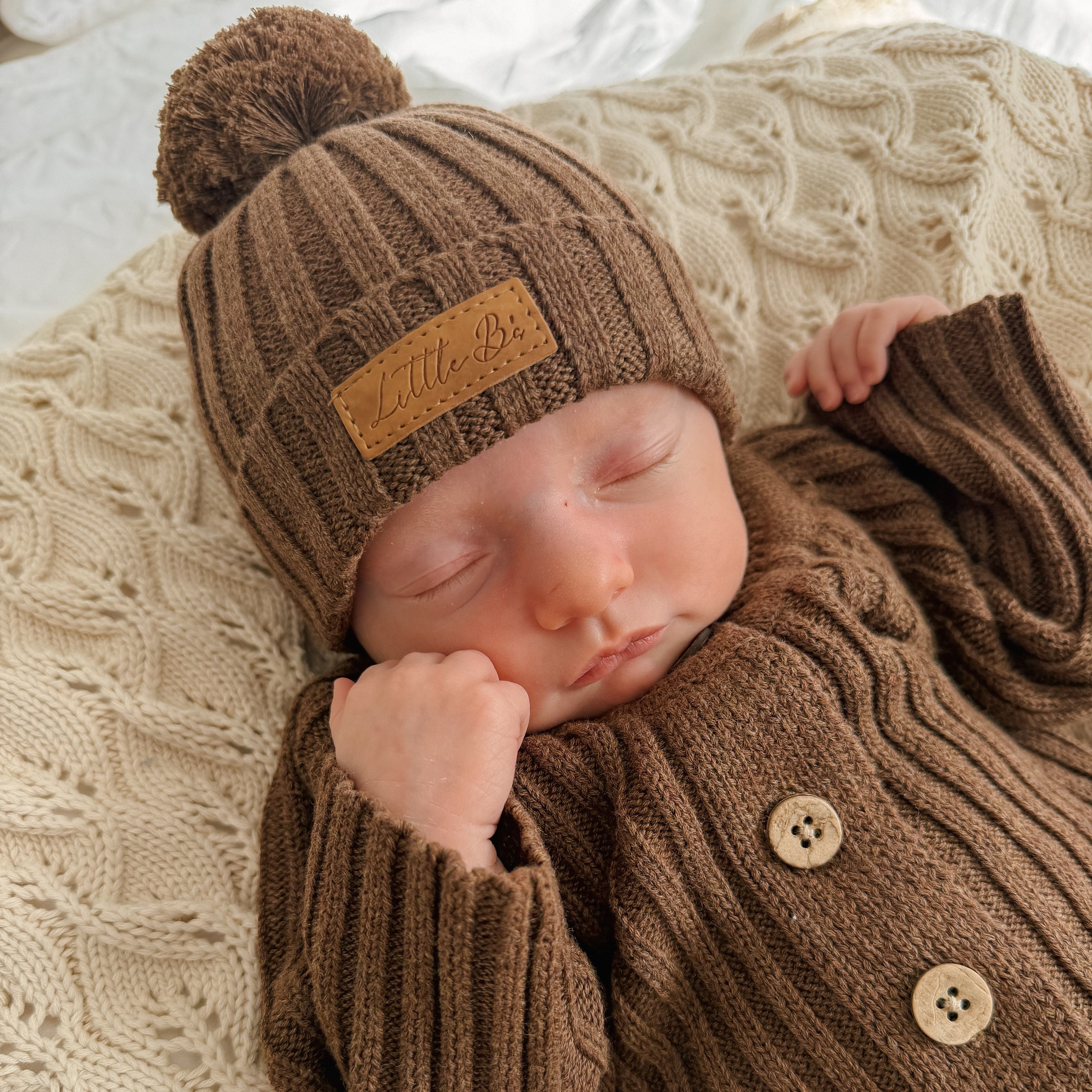 Baby wearing knitted ribbed chocolate romper and matching beanie on beige knitted blanket 