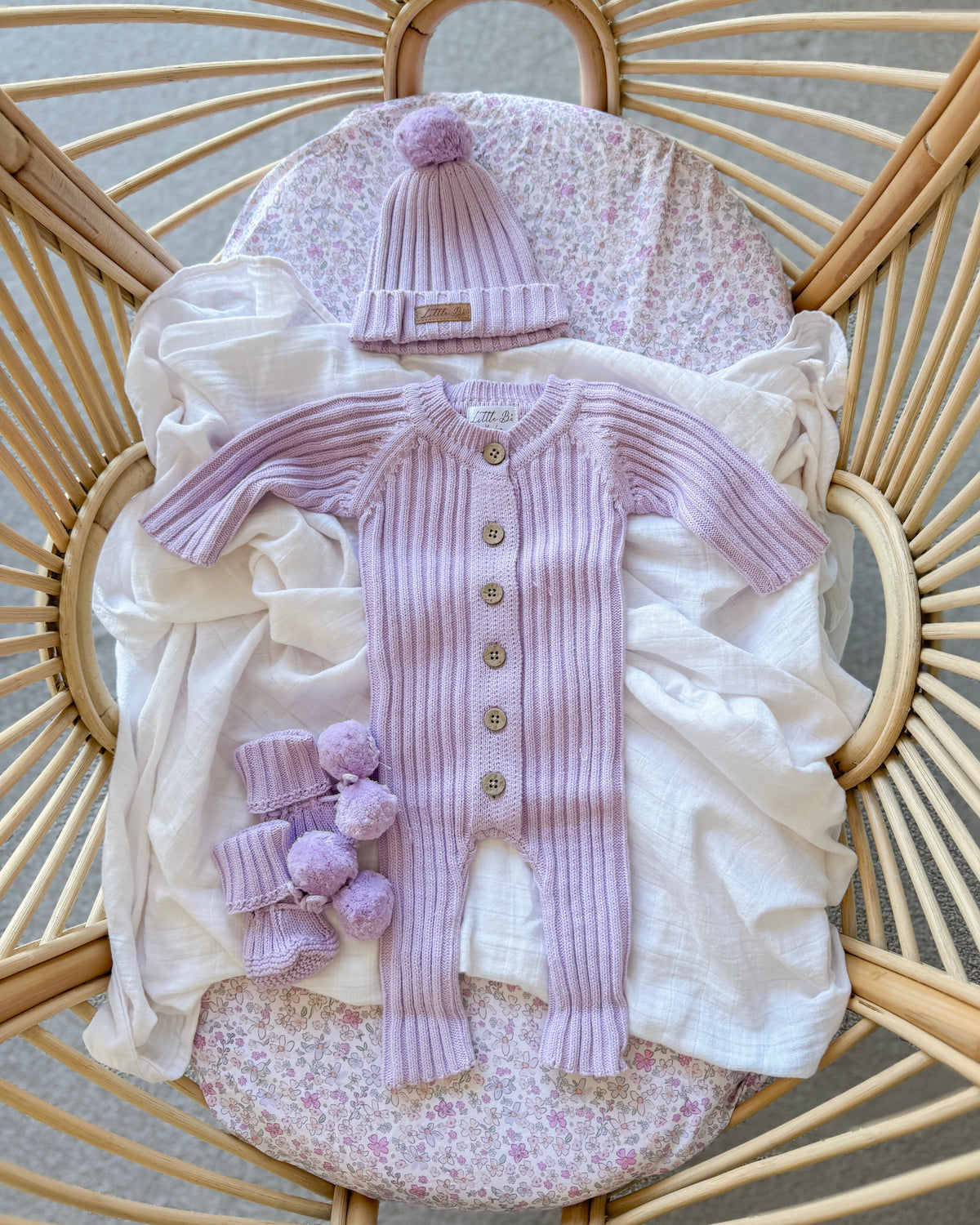 A baby clothing set in soft lavender and white, including a ribbed one-piece with front buttons, a matching pom-pom beanie, and matching leg warmers, arranged on a floral cushion inside a wicker chair.
