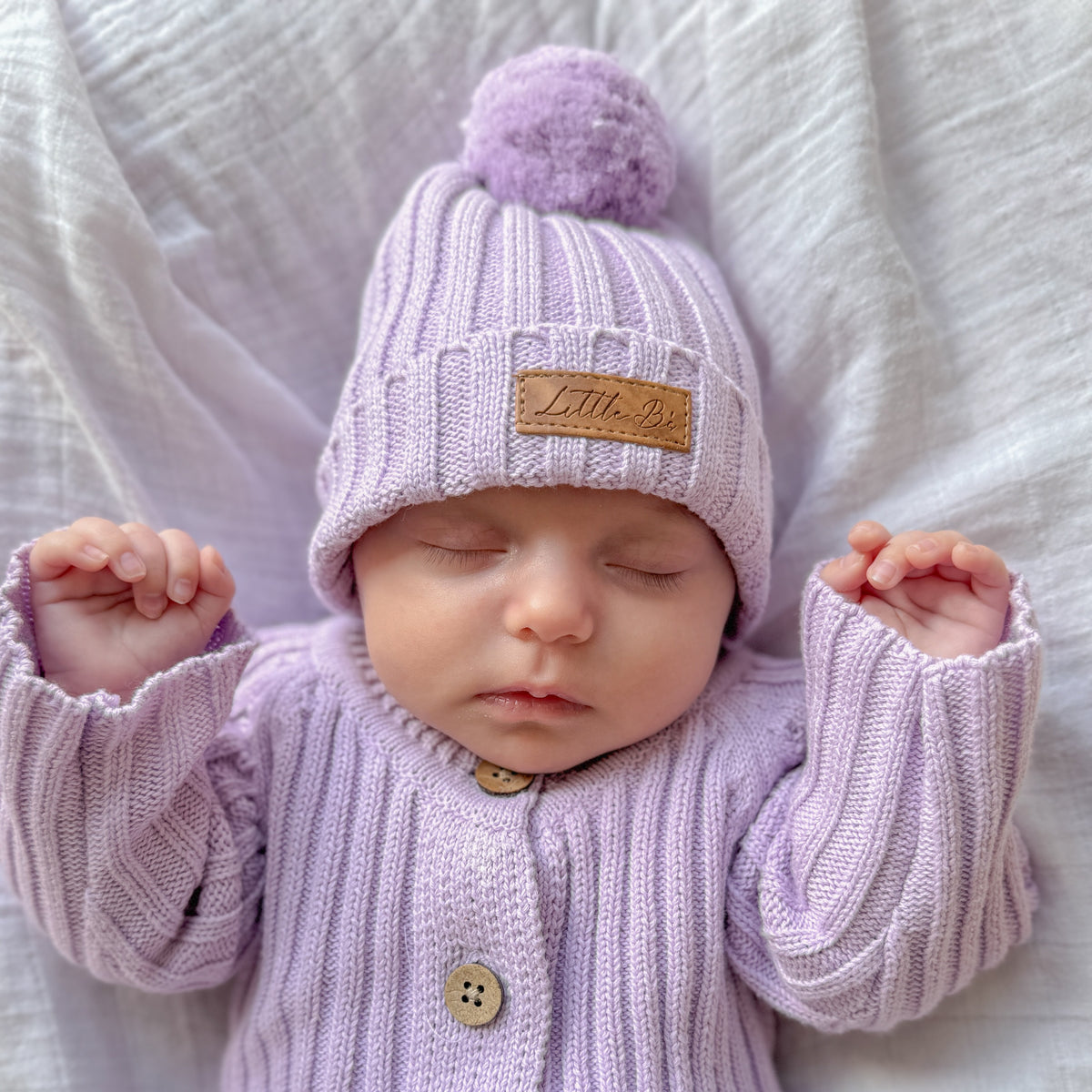 A sleeping newborn baby wearing a light purple ribbed knit cardigan and matching pom-pom beanie with a tan leather patch that reads 'Little Be'. The baby is lying on a textured white blanket.
