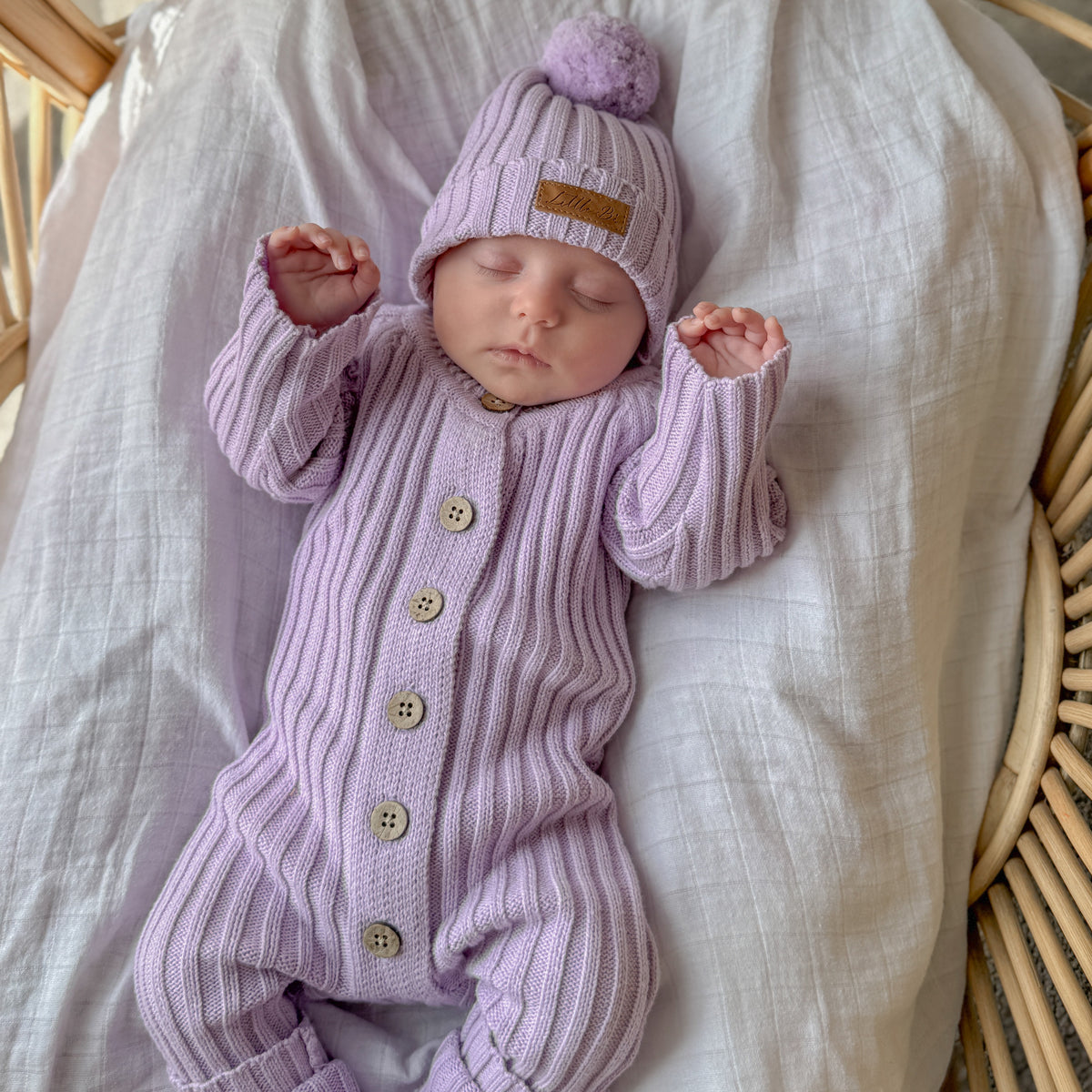 A sleeping newborn baby wearing a light purple ribbed knit one-piece jumpsuit and matching pom-pom beanie, resting in a woven rattan bassinet on a white muslin blanket.