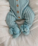 A baby wearing a blue ribbed romper suit with vintage wooden buttons, paired with a matching blue booties with pom poms and drawstring laying on a cozy white blanket.