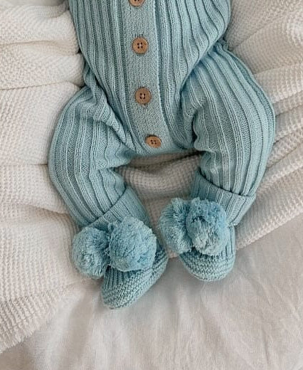 A baby wearing a blue ribbed romper suit with vintage wooden buttons, paired with a matching blue booties with pom poms and drawstring laying on a cozy white blanket.