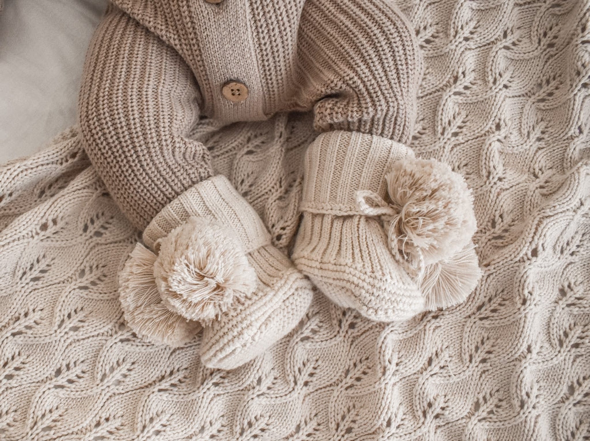 A baby wearing a pair of beige knitted booties with pom pom details, displayed on a textured knitted blanket.