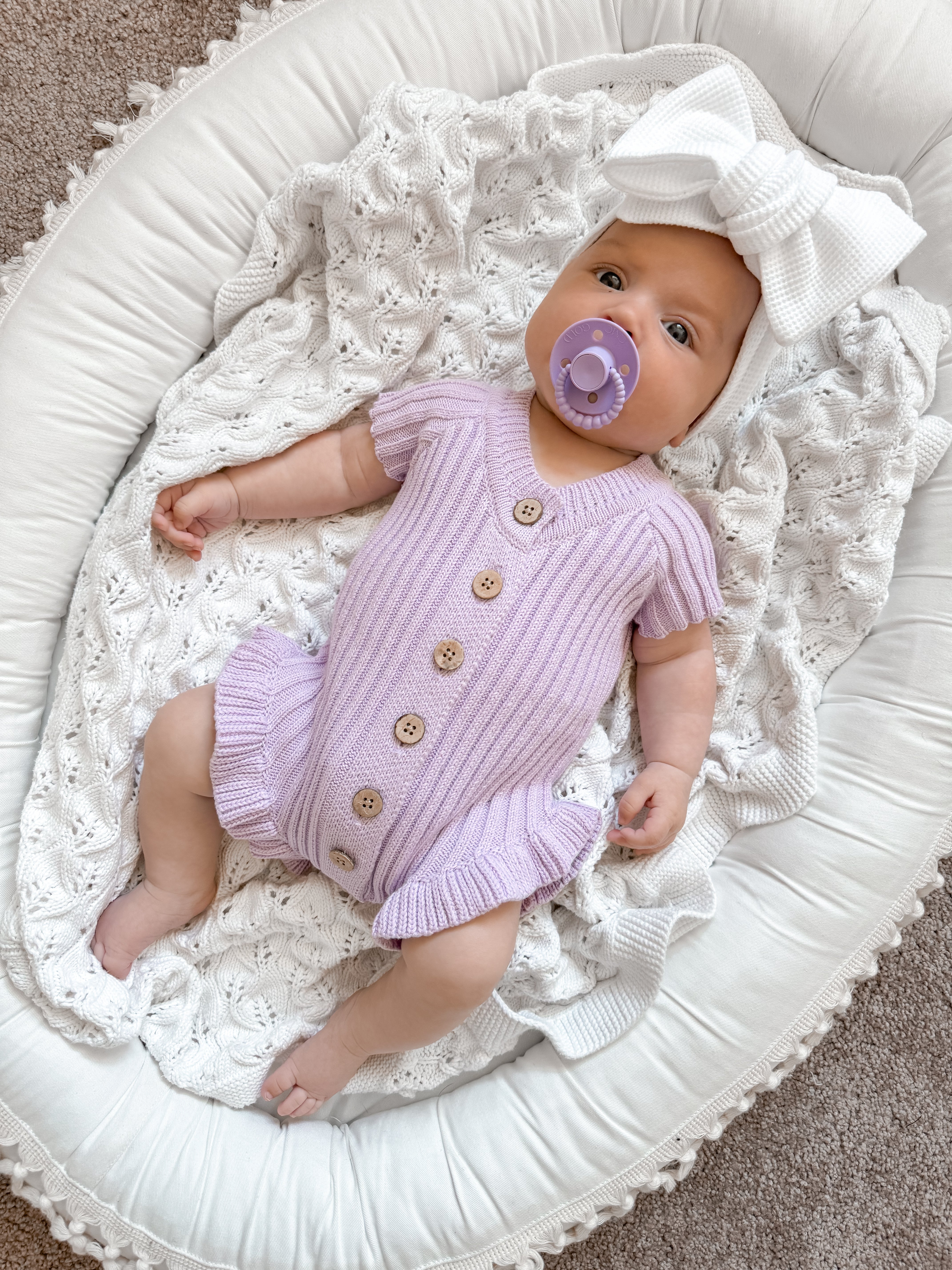 Little B's Knitted Cotton Ribbed Ruffle Romper in purple on a baby with a white bow and pacifier