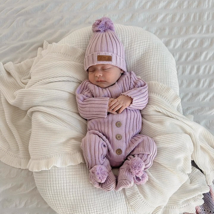 A sleeping newborn baby wearing a matching light purple knit hat with a pom-pom and a ribbed purple one-piece romper with button details, lying on a textured white bassinet surrounded by soft cream blankets.