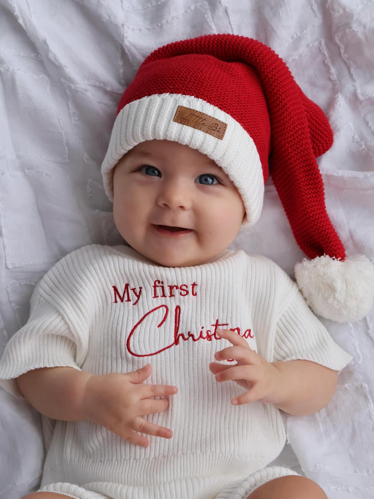 A smiling baby lying on a white textured blanket, wearing a red and white knit Santa hat with a white pom-pom and a white ribbed bodysuit with red text reading 'My first Christmas'.