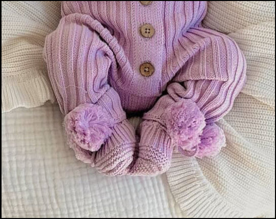 A new born baby wearing cotton purple knitted booties with pom poms and a ribbed beanie, lying on a cushioned baby nest.