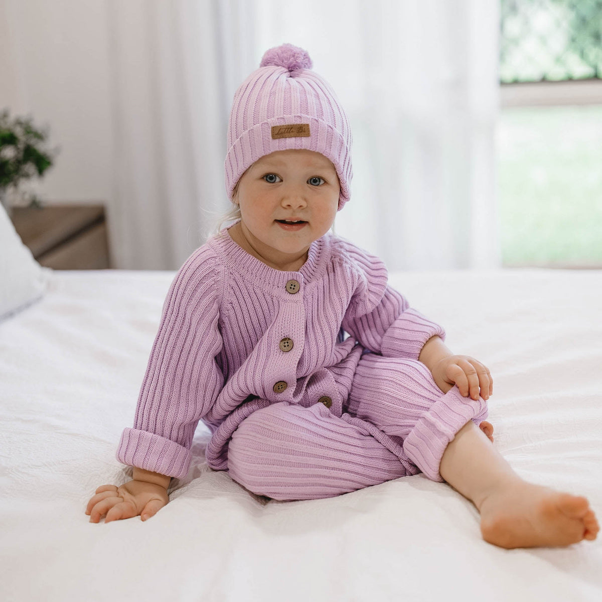A baby sitting on a white bed wearing a matching light purple ribbed knit romper, buttoned up jacket, and pom-pom beanie.