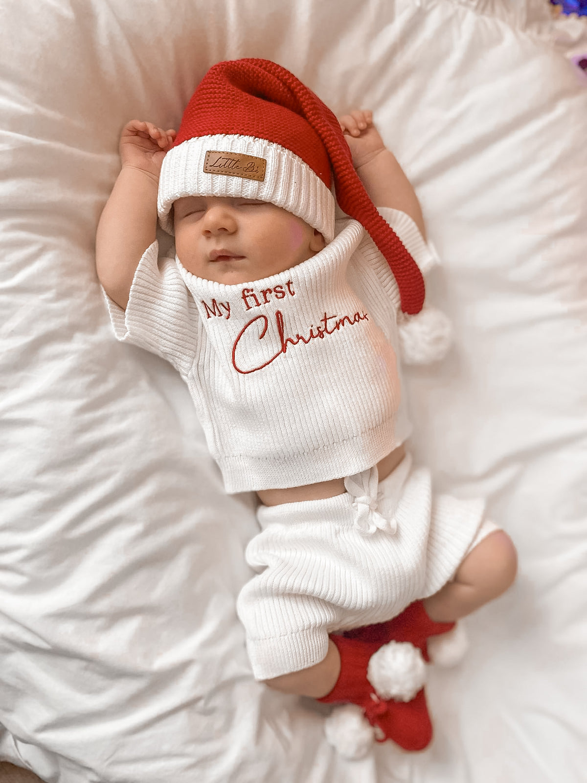A sleeping newborn baby lying on white bedding, wearing a white ribbed knit outfit with 'My First Christmas' embroidered in red on the chest, a red Santa hat with white pom-poms, and red booties with white pom-poms.
