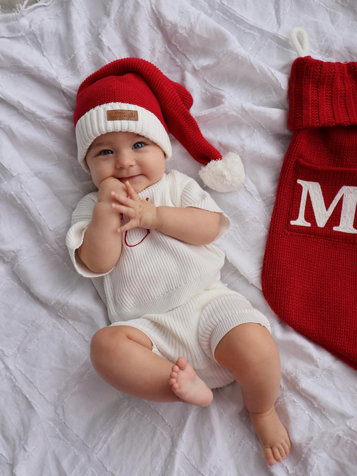 A baby lying on white textured bedding, wearing a red and white knit Santa hat and a white ribbed short-sleeve romper. A red knit stocking with a white letter 'M' is partially visible on the right.