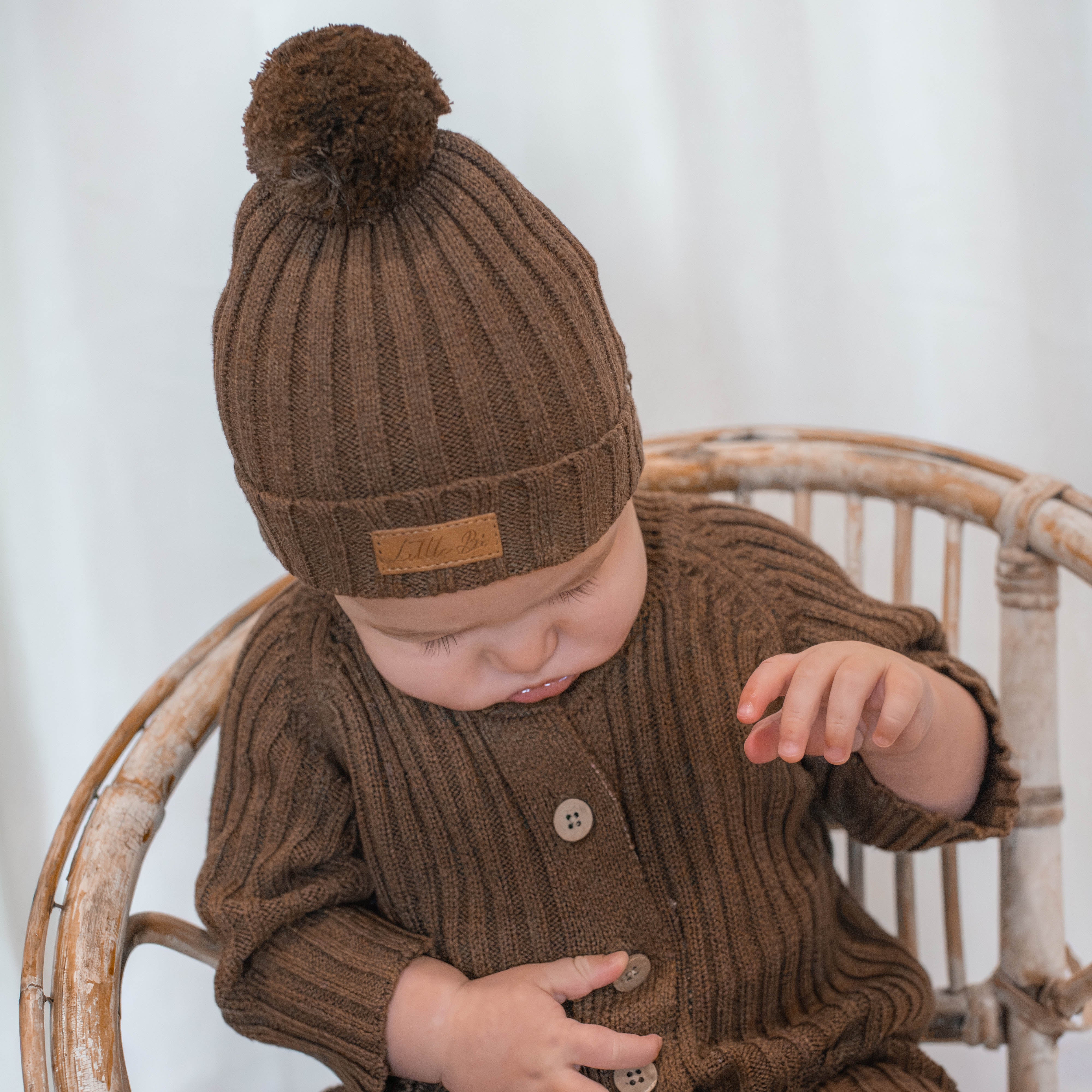 Baby sitting in cane chair wearing chocolate ribbed knitted beanie and matching romper