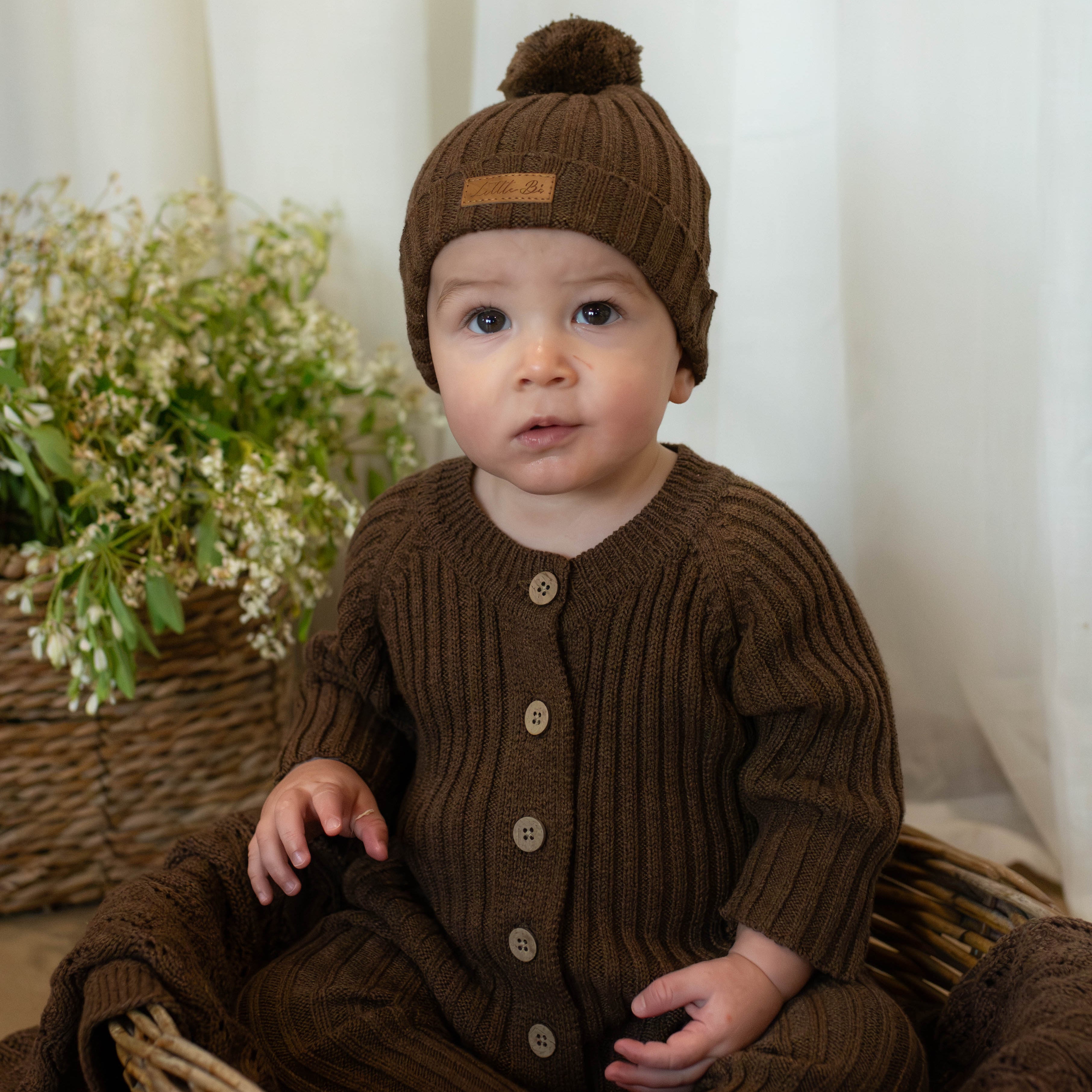 8 month old baby sitting in cane basket wearing knitted brown ribbed romper and beanie 