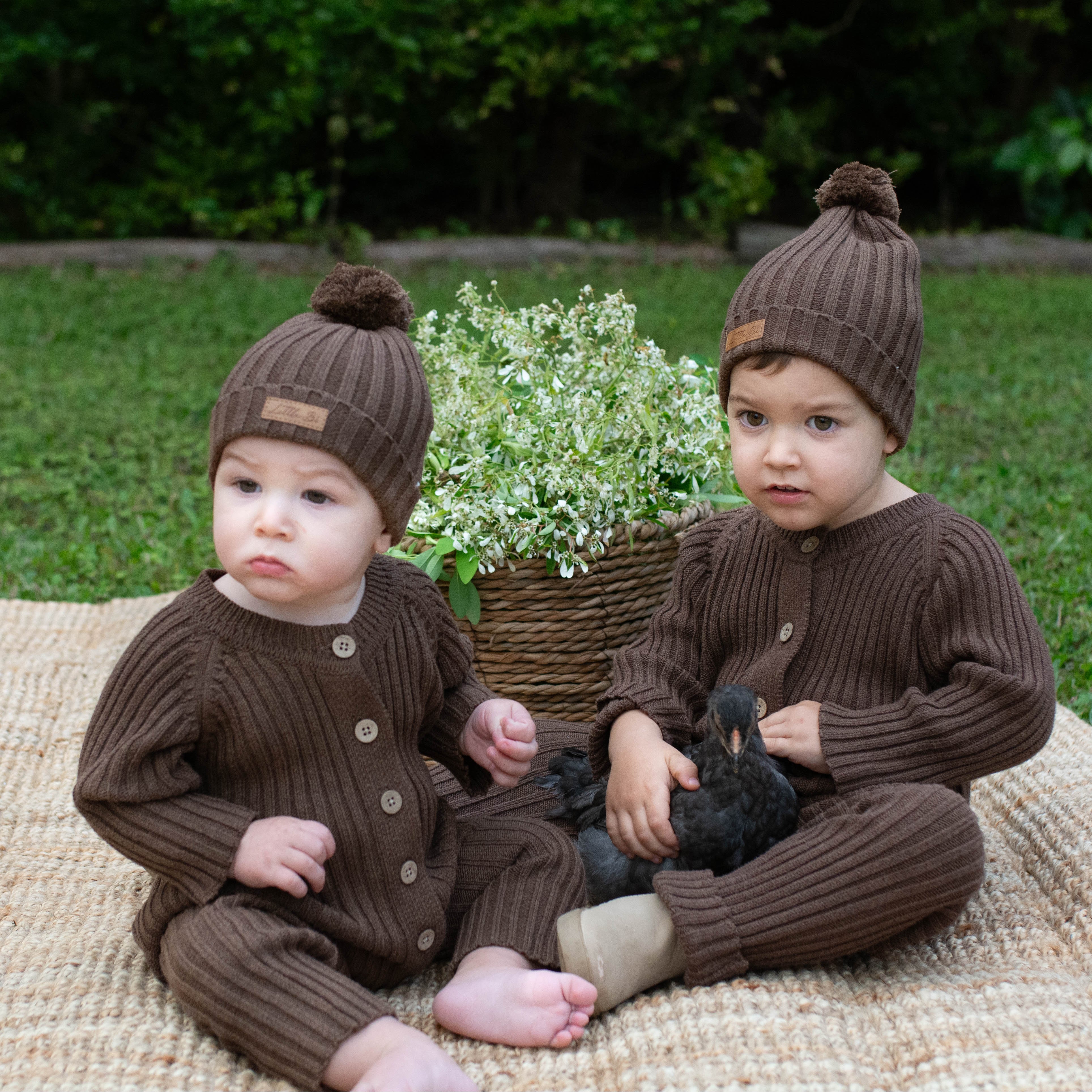 Two young boys sitting on a mat outdoors holding a young black chicken wearing brown knitted ribbed rompers.