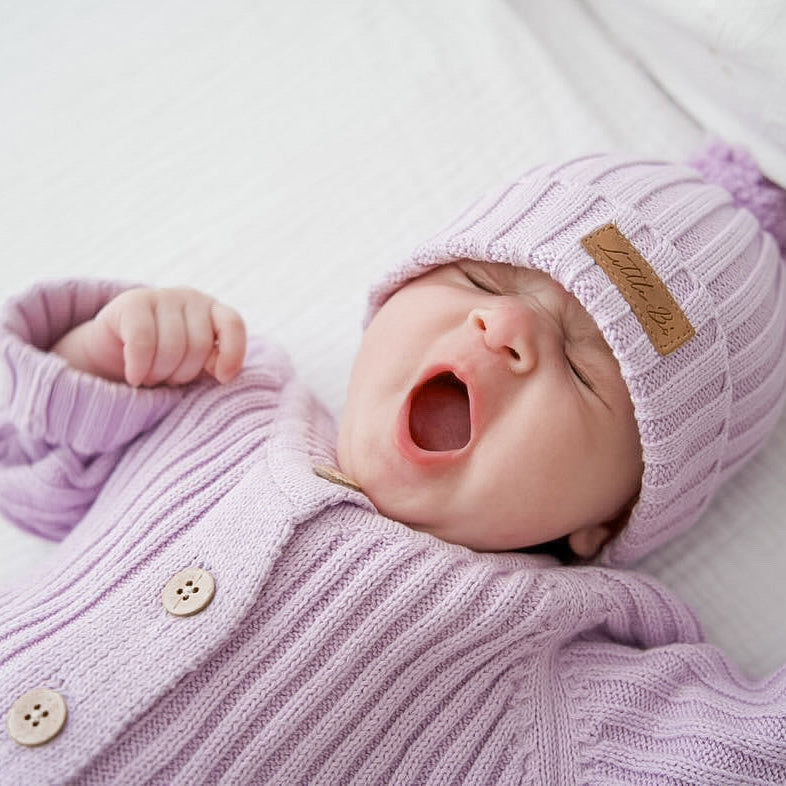 A newborn baby wearing a light purple ribbed knit cardigan and matching pom-pom beanie with a small tan brand patch, yawning while lying on a white quilted blanket.