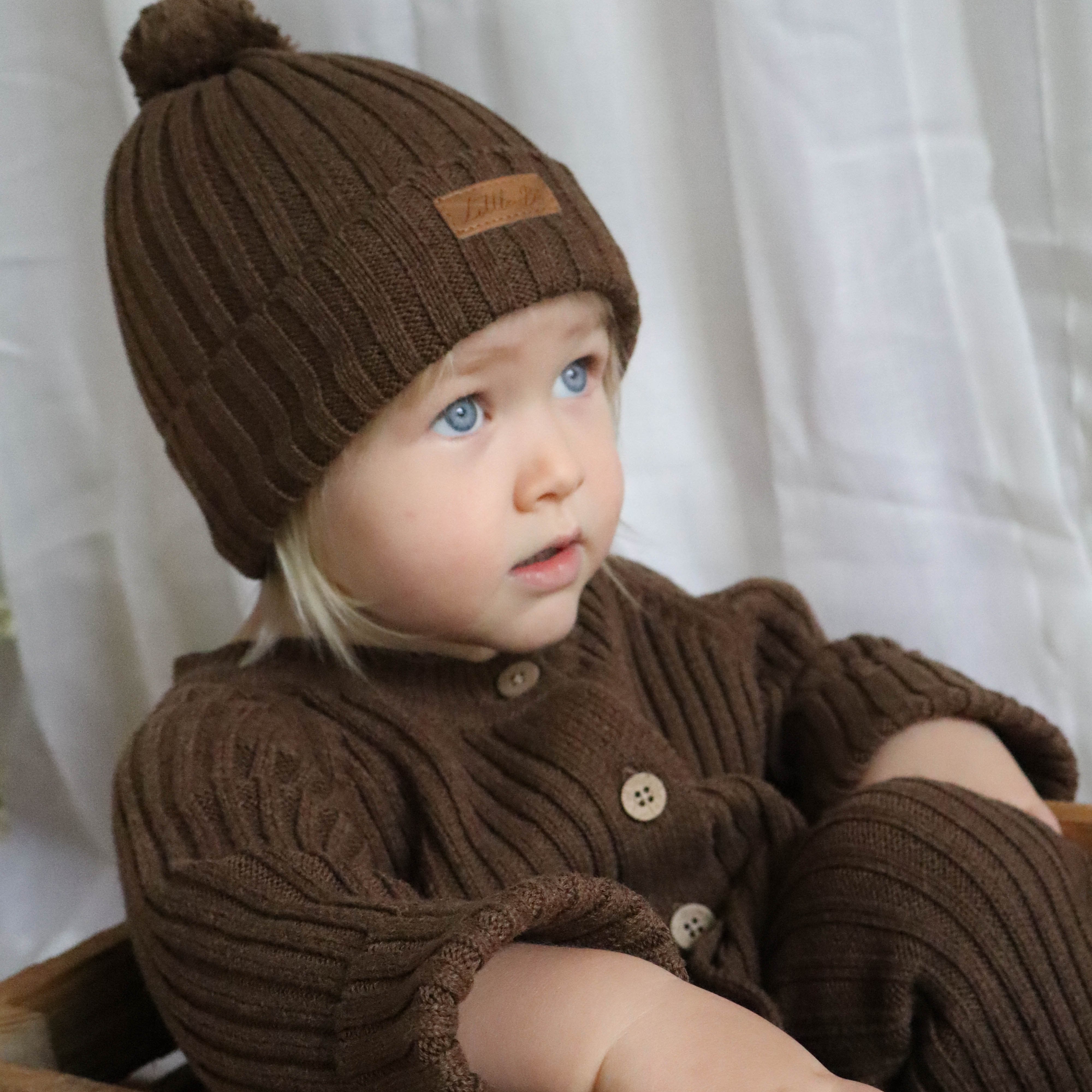 Young girl sitting in a timber crate wearing a chocolate knitted ribbed romper and matching beanie with white background