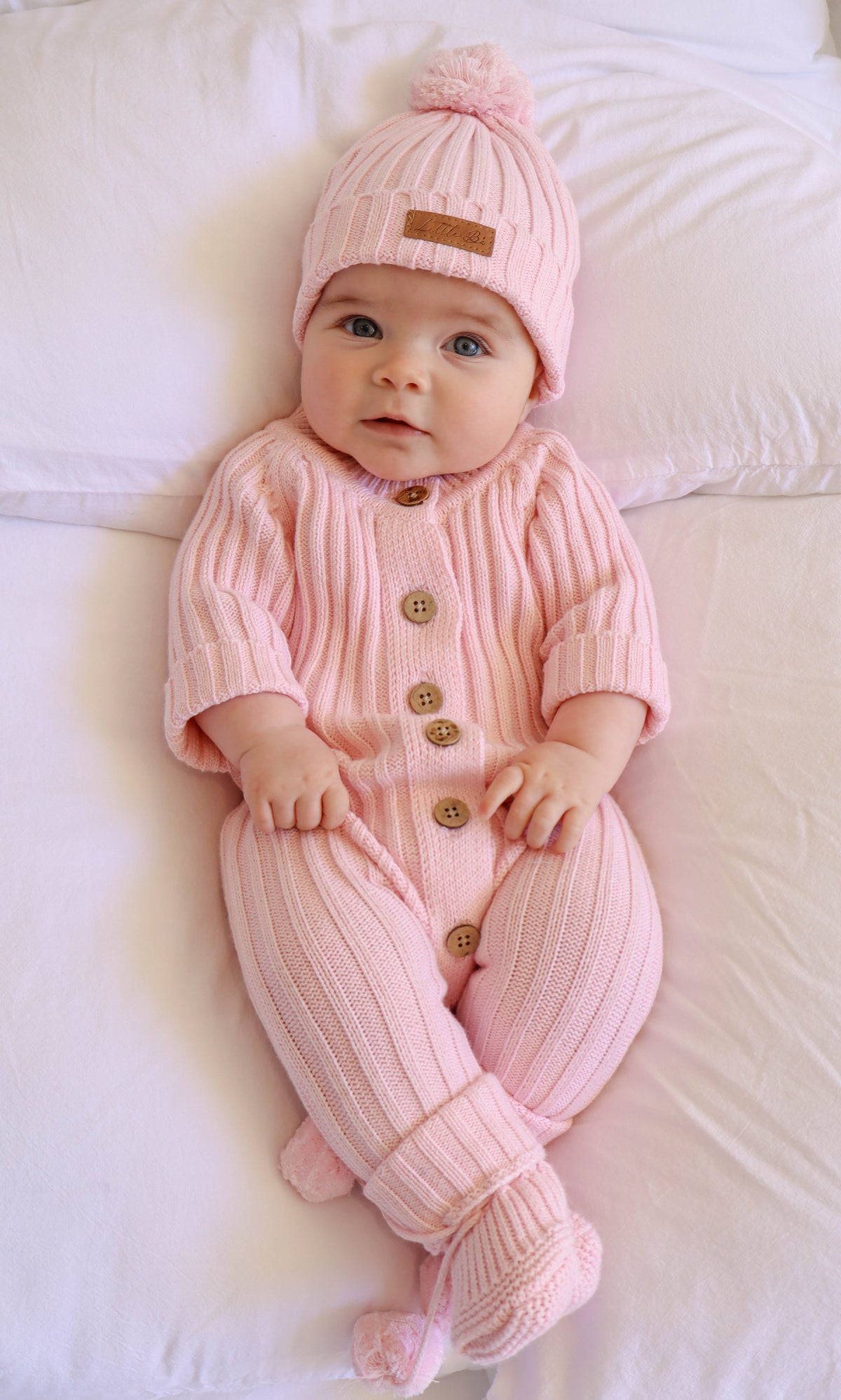 A baby lying on white bedding wearing a pink ribbed knit one-piece jumpsuit with button closures and a matching pink knit pom-pom hat.