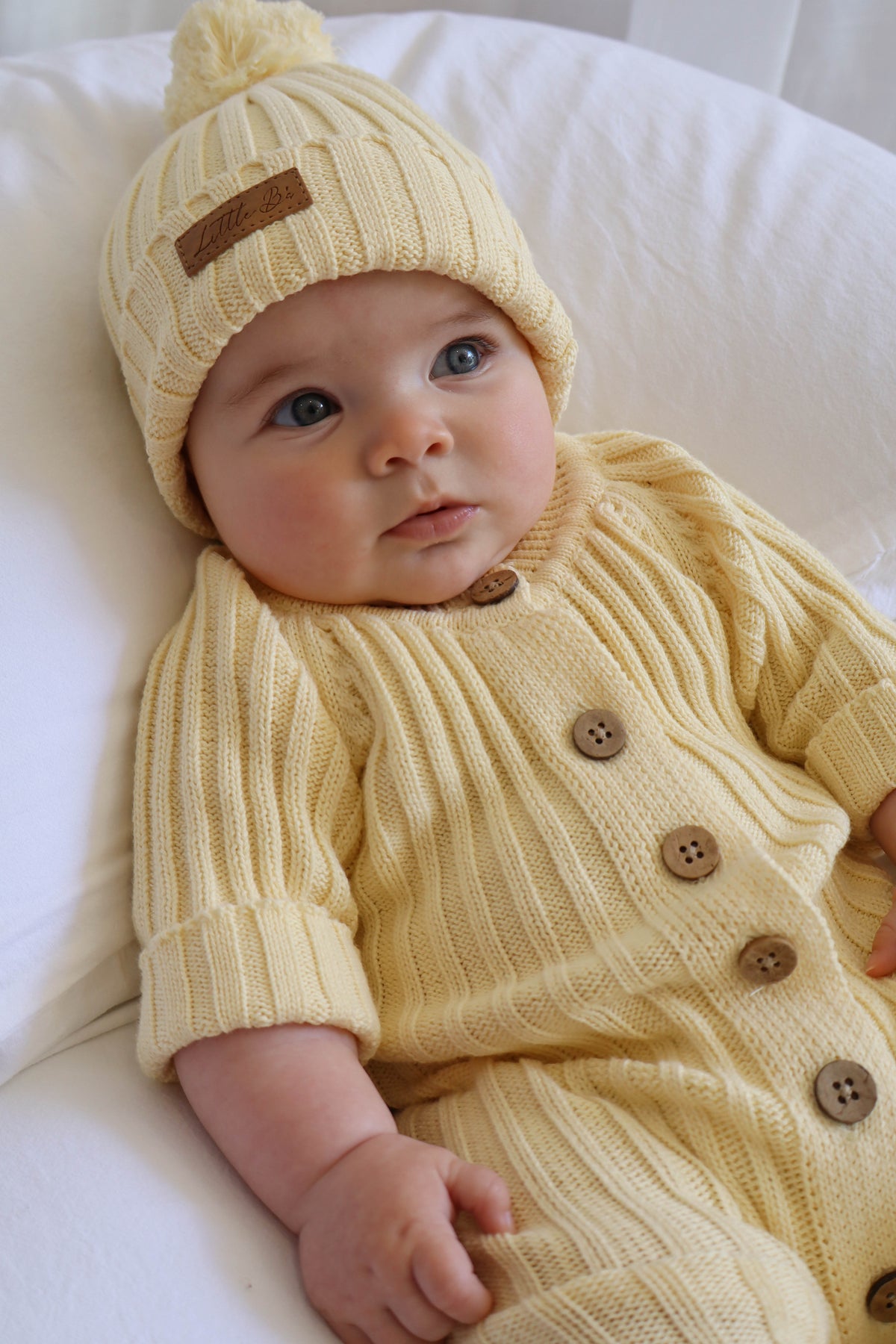 A baby lying on a white cushion, wearing a pale yellow ribbed-knit bonnet with a pom-pom and a matching long-sleeve button-front romper. The knit outfit has brown wooden buttons and a small brown patch on the bonnet.