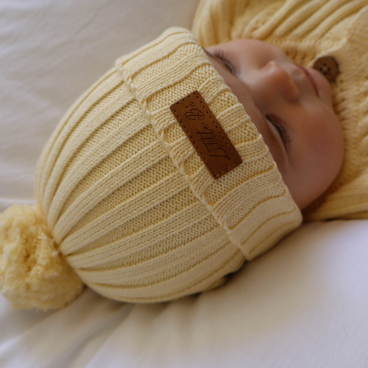 A baby wearing a cream ribbed knit pom-pom beanie and a matching cream cardigan, lying on a white blanket. The beanie features a brown faux-leather patch with the brand name 'Bella+Canvas' visible.