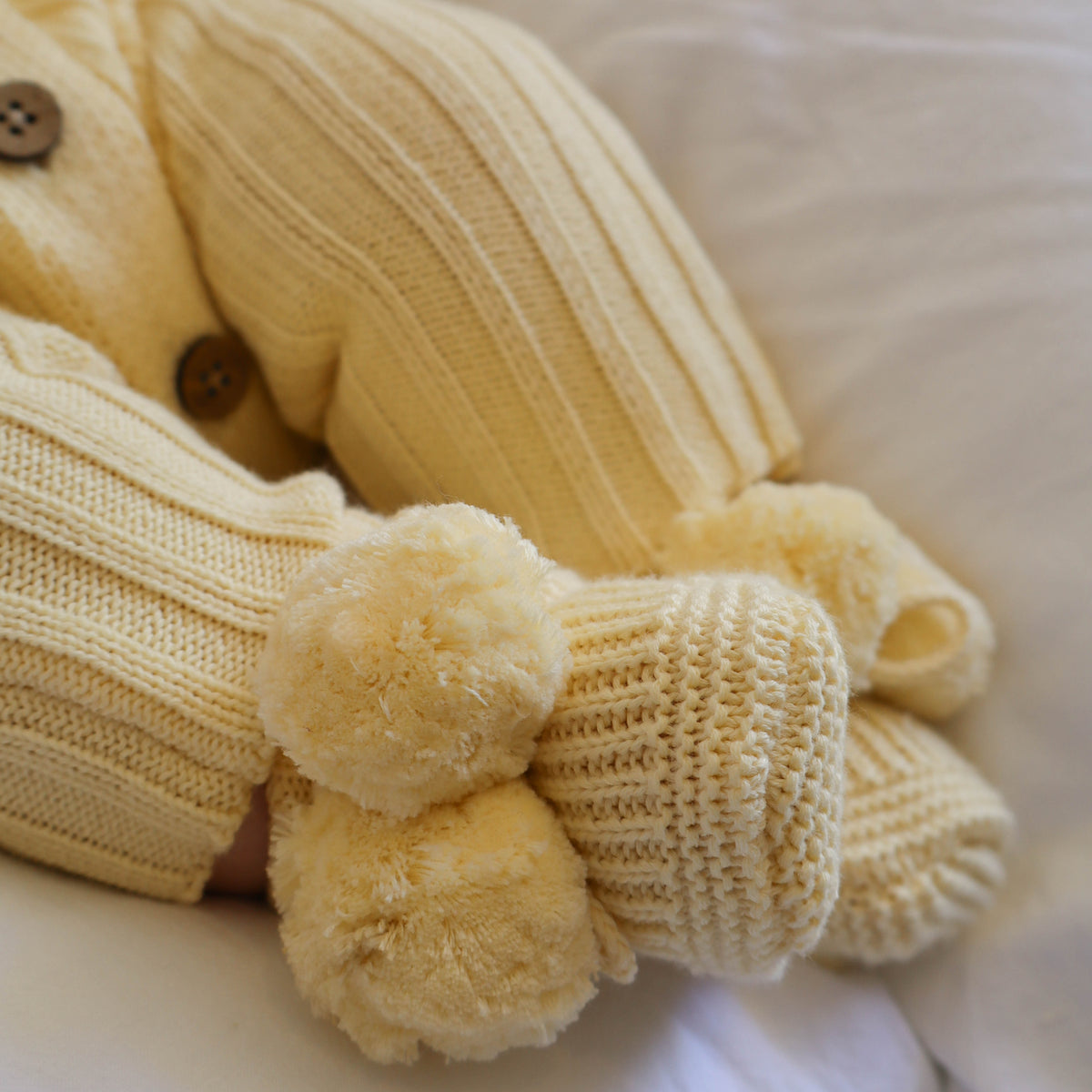 A close-up of a pale yellow ribbed knit pom pom bootie with matching knit romper and fluffy pom-pom details, resting on a soft white fabric background.