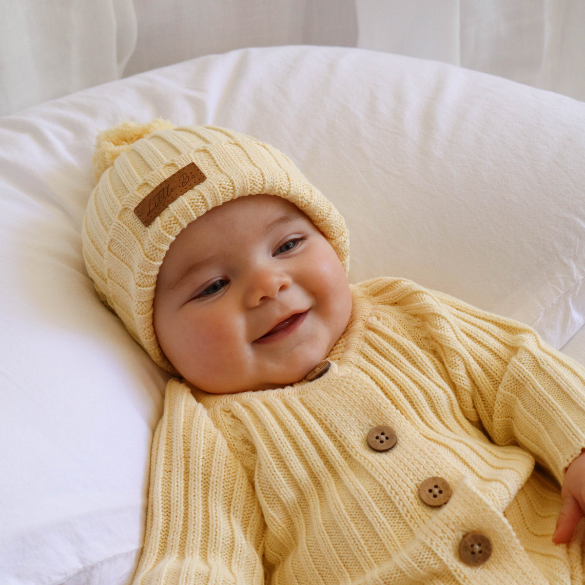 A baby wearing a yellow-coloured ribbed knit bonnet with a brown patch and a matching yellow ribbed knit romper with brown buttons, lying on a white pillow.