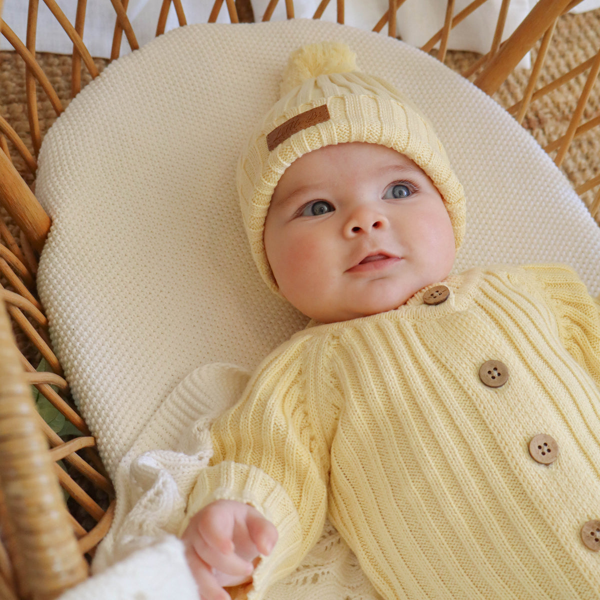 A baby lying on a cream cushion inside a wicker bassinet, wearing a pale yellow ribbed knit bonnet and matching long-sleeve button-front romper.