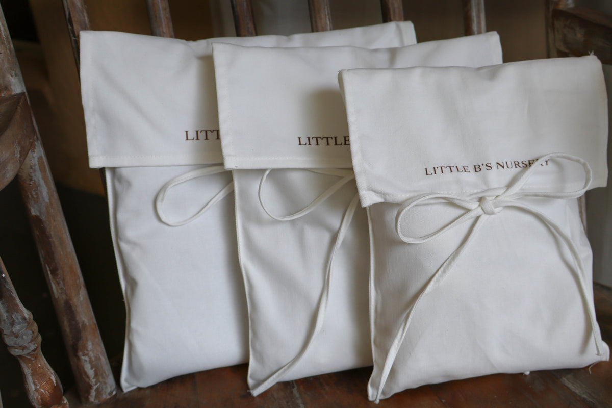 Three cream-colored cotton canvas pouches with drawstring ties and embroidered text, displayed on a rustic wooden chair.