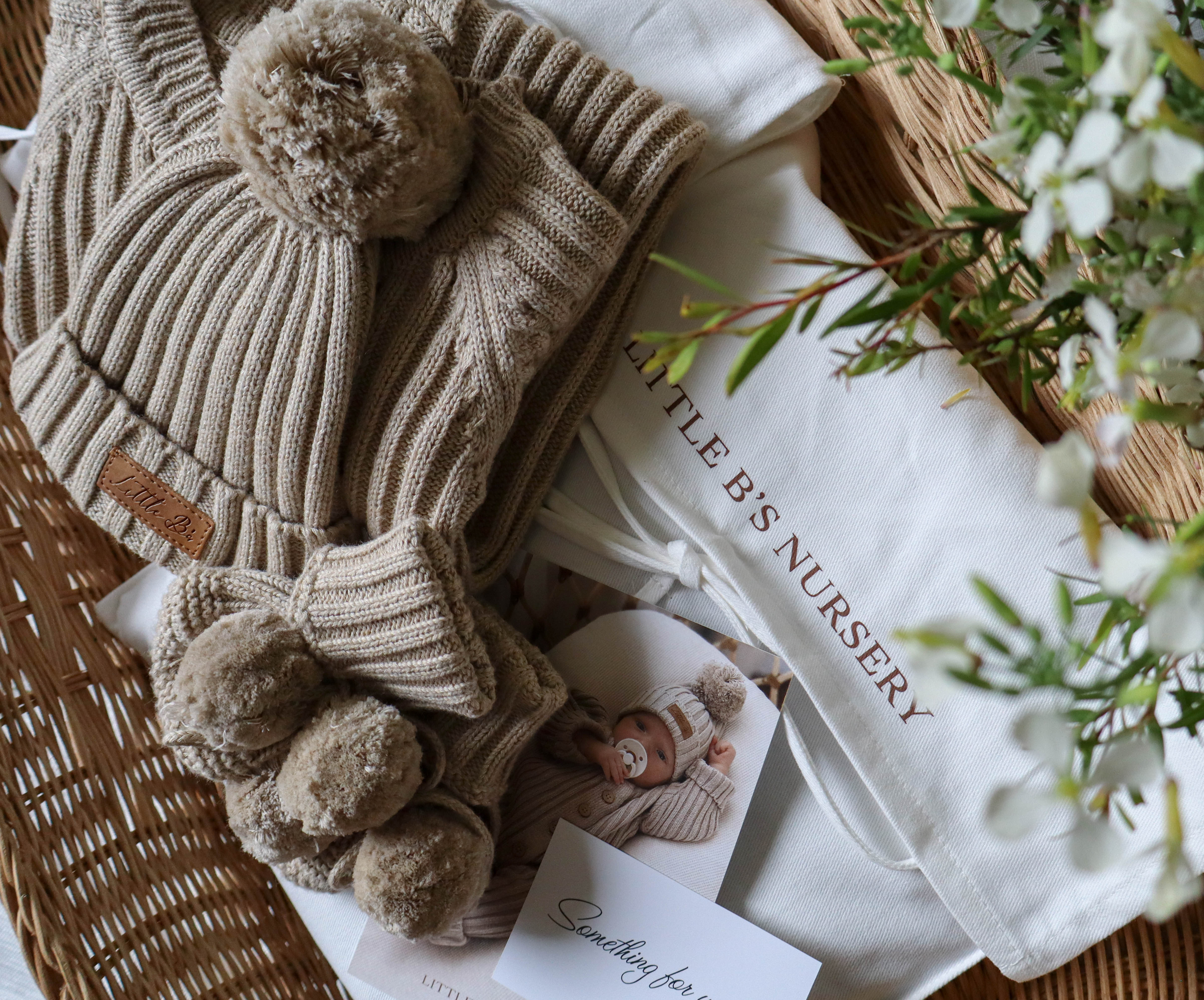 A styled flat-lay of a baby knit hat and matching mittens in a warm beige/tan color, arranged in a wicker basket with a white drawstring pouch printed with 'MILLE BIS NURSERY' and baby photos.
