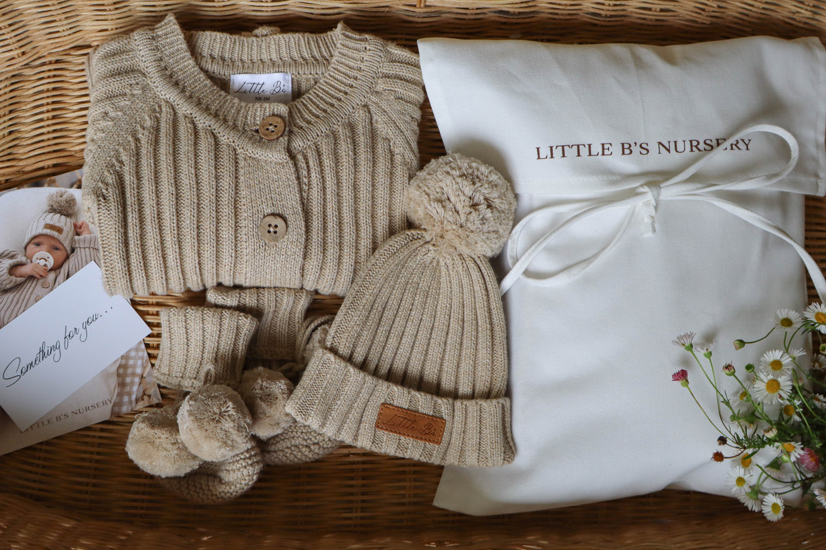 A baby knit outfit set in a wicker basket, featuring a beige ribbed cardigan, matching hat with pom-poms, and booties, alongside a white 'LITTLE B'S NURSERY' gift bag and a small card.
