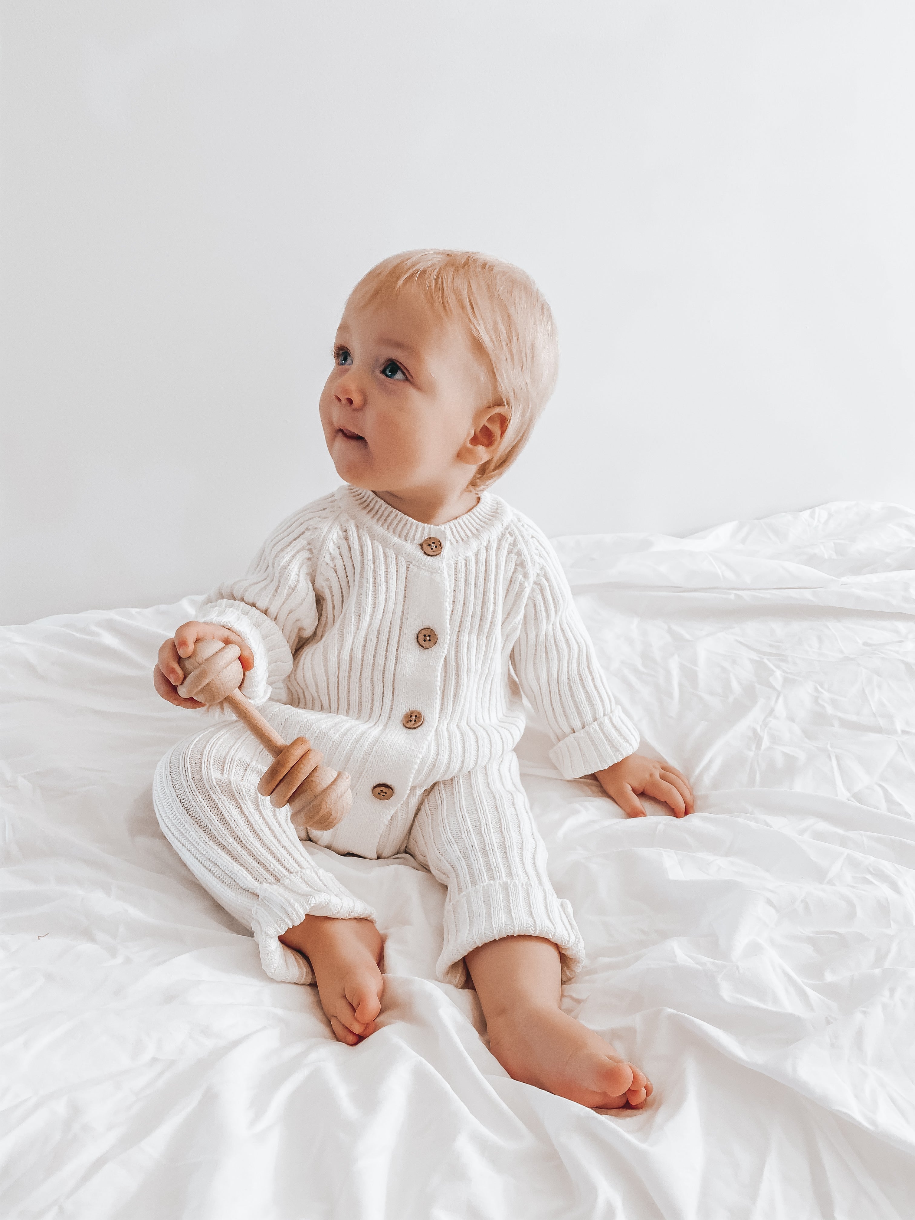 Little B's Retail Knitted Cotton Ribbed Romper - Milk, worn by a baby holding a wooden rattle