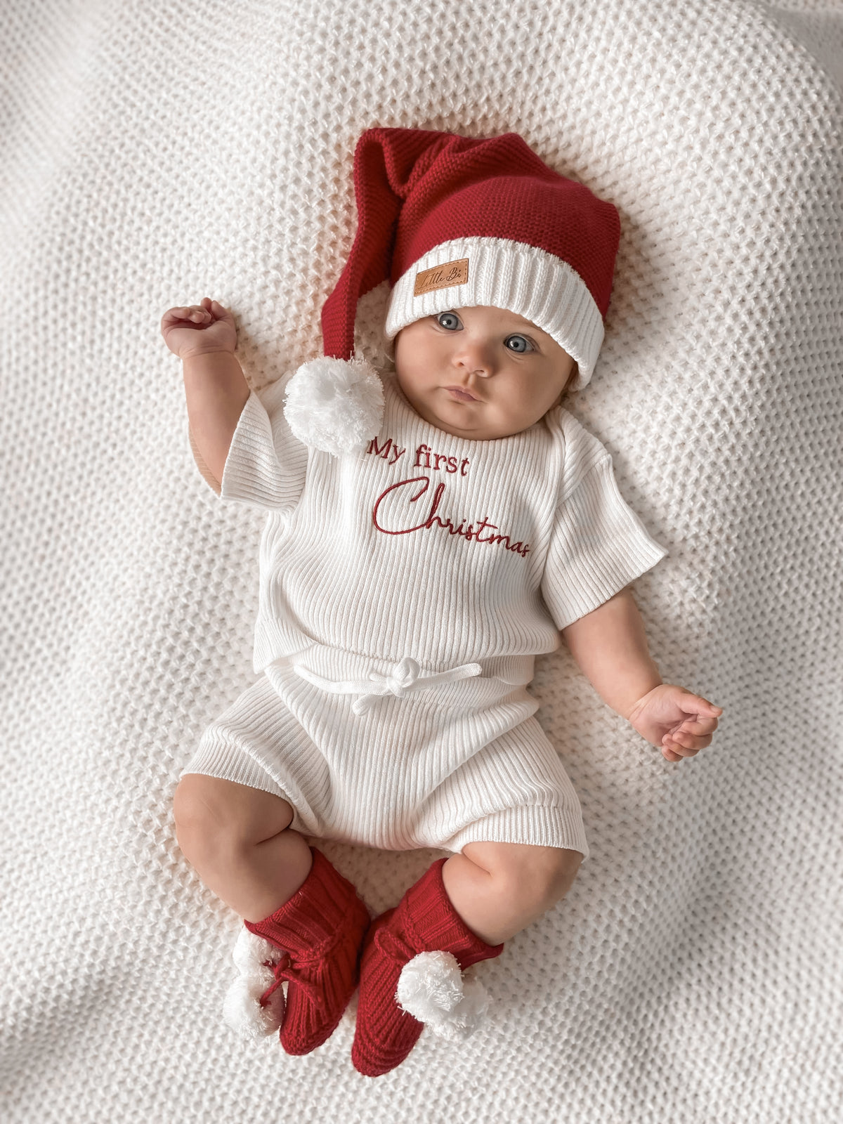 A baby lying on a textured cream blanket, wearing a red and white Santa hat, a white ribbed short-sleeve romper with 'My First Christmas' embroidered in red, and red booties with white pom-poms.